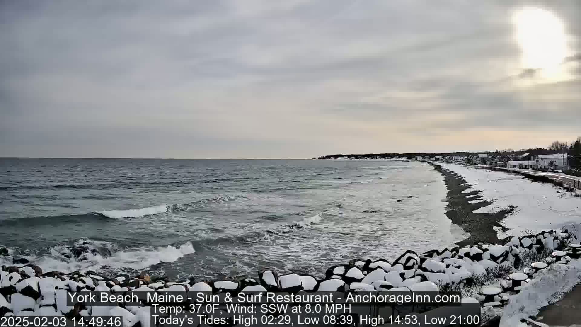 A partially sunny, cold day shows a snow-covered beach with a dark, rocky shoreline and ocean waves.
