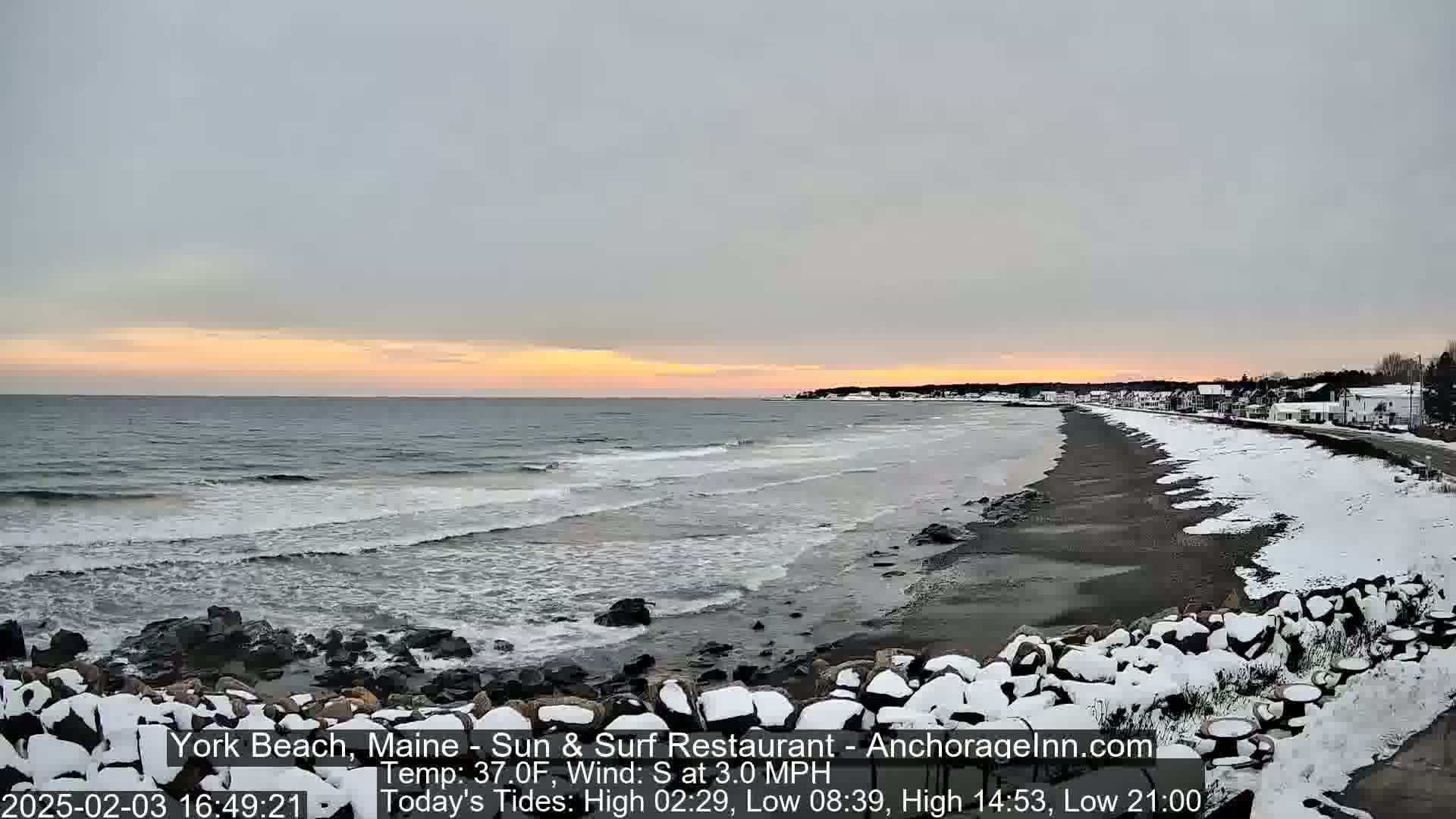 A snow-dusted beach with dark rocks and a calm ocean under a pale sunset sky.
