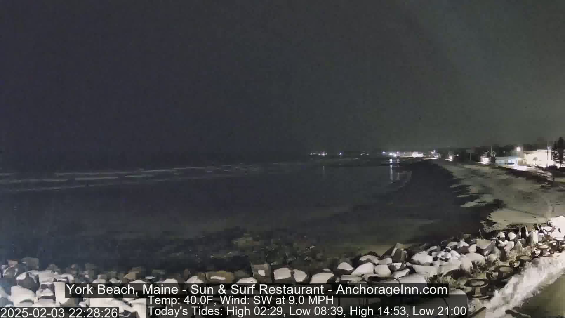 A nighttime view of a dark, snow-dusted beach with a rocky shoreline and gently rolling waves under a cloudy sky.