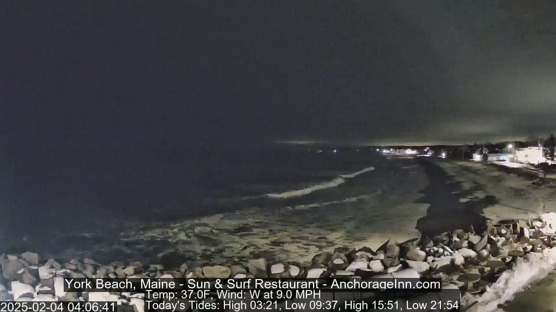 A nighttime view of a rocky shoreline with dark, wavy water under a cloudy sky.