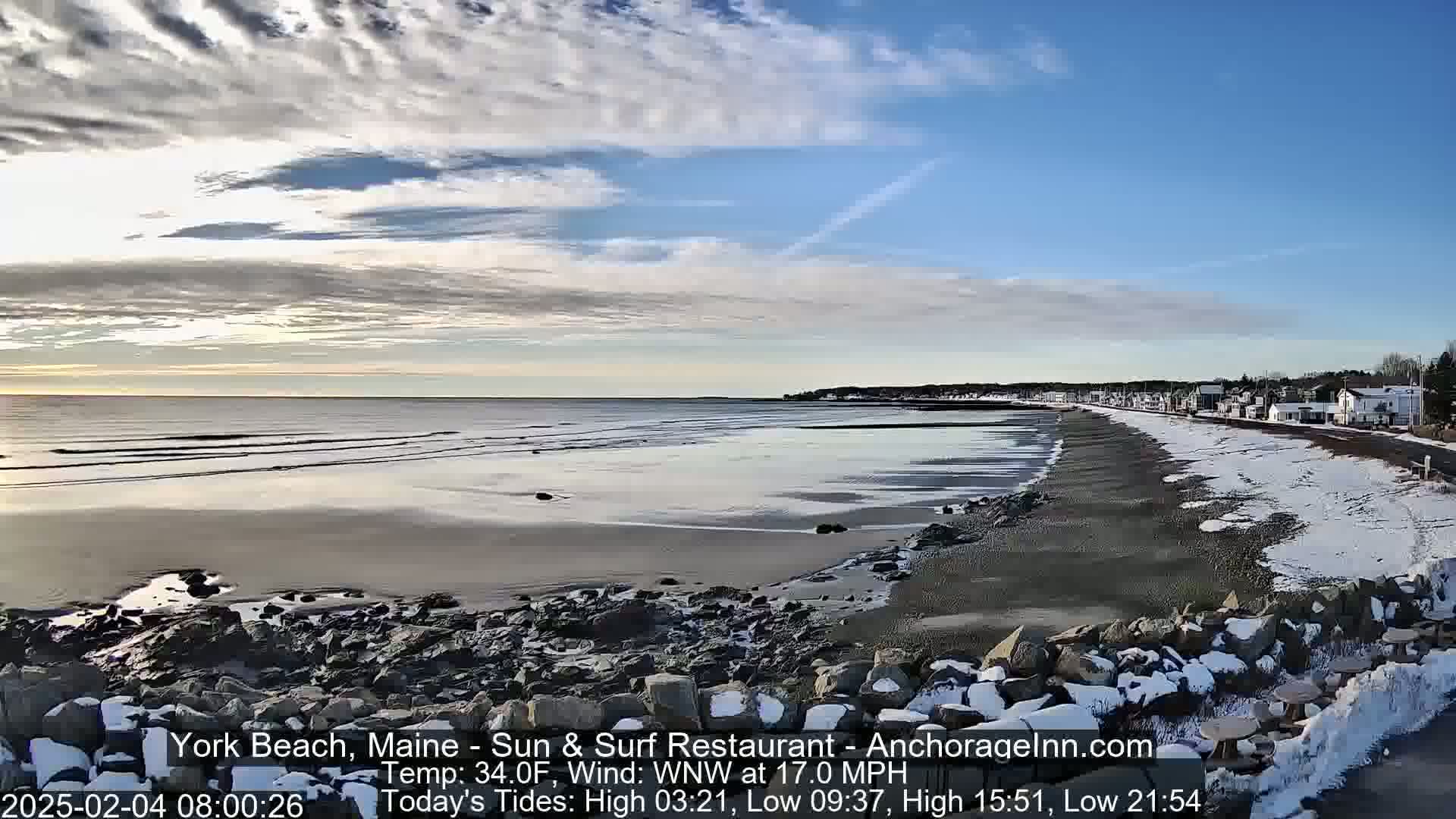 A partly cloudy, cold day at a snowy beach with a calm ocean, showing a shoreline with buildings in the distance.