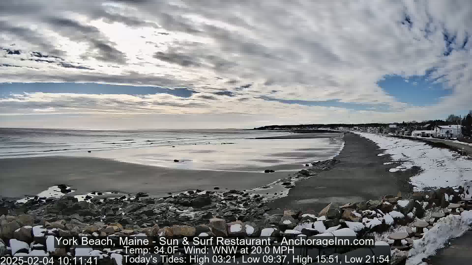 A partially snowy beach stretches along a calm ocean under a mostly cloudy sky.