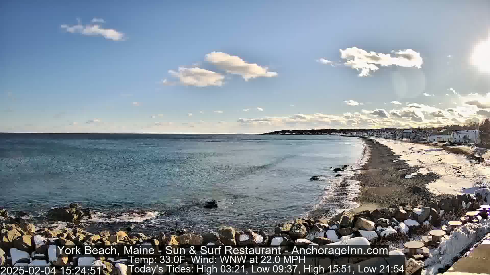 A partly cloudy day with a 22 mph wind shows a rocky coastline with a sandy beach partially covered in snow and a calm ocean.