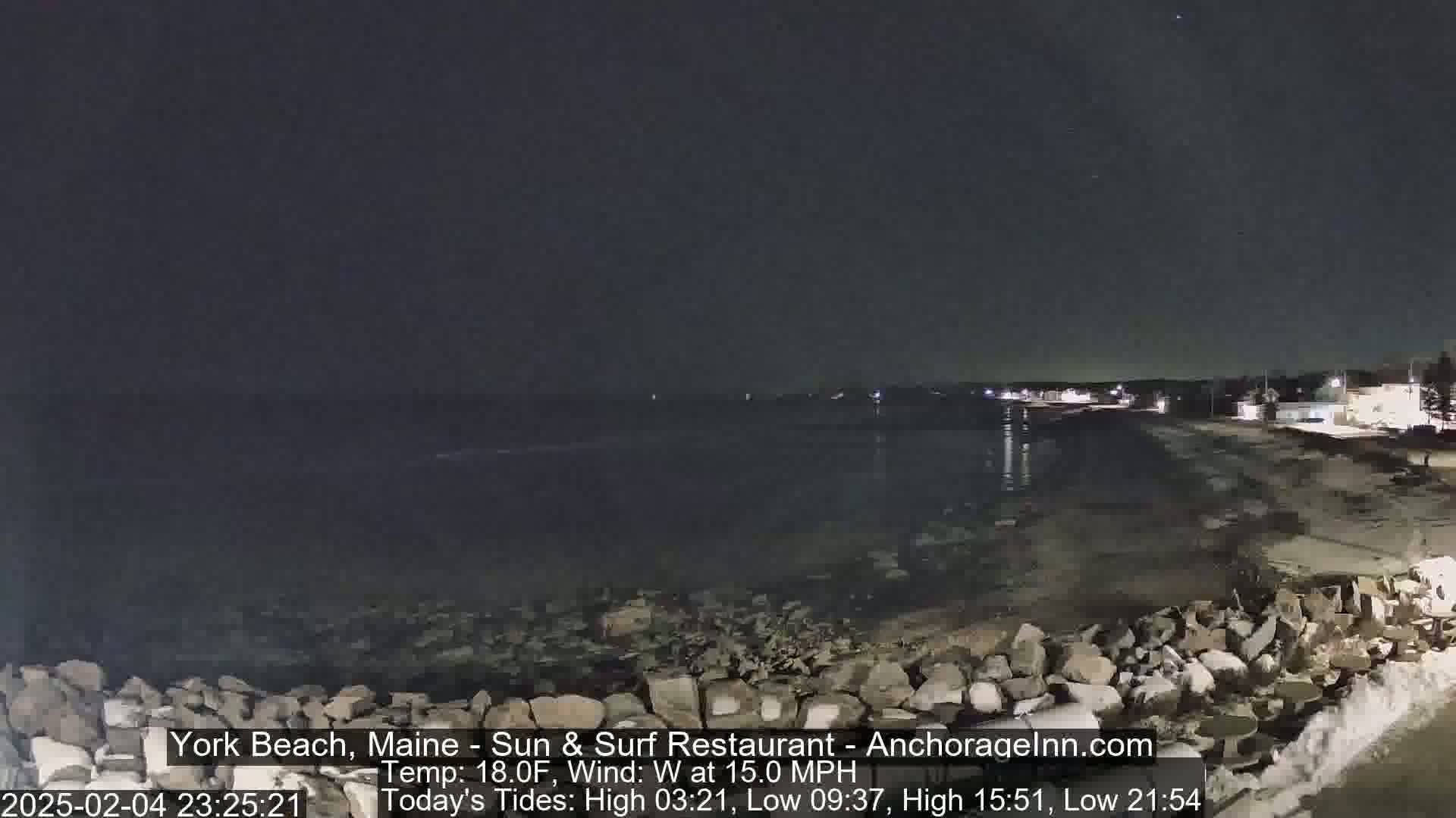 A nighttime coastal scene shows a dark ocean, a sandy beach, and a rocky shoreline under a clear, dark sky with a 15 mph westerly wind and 18°F temperature.