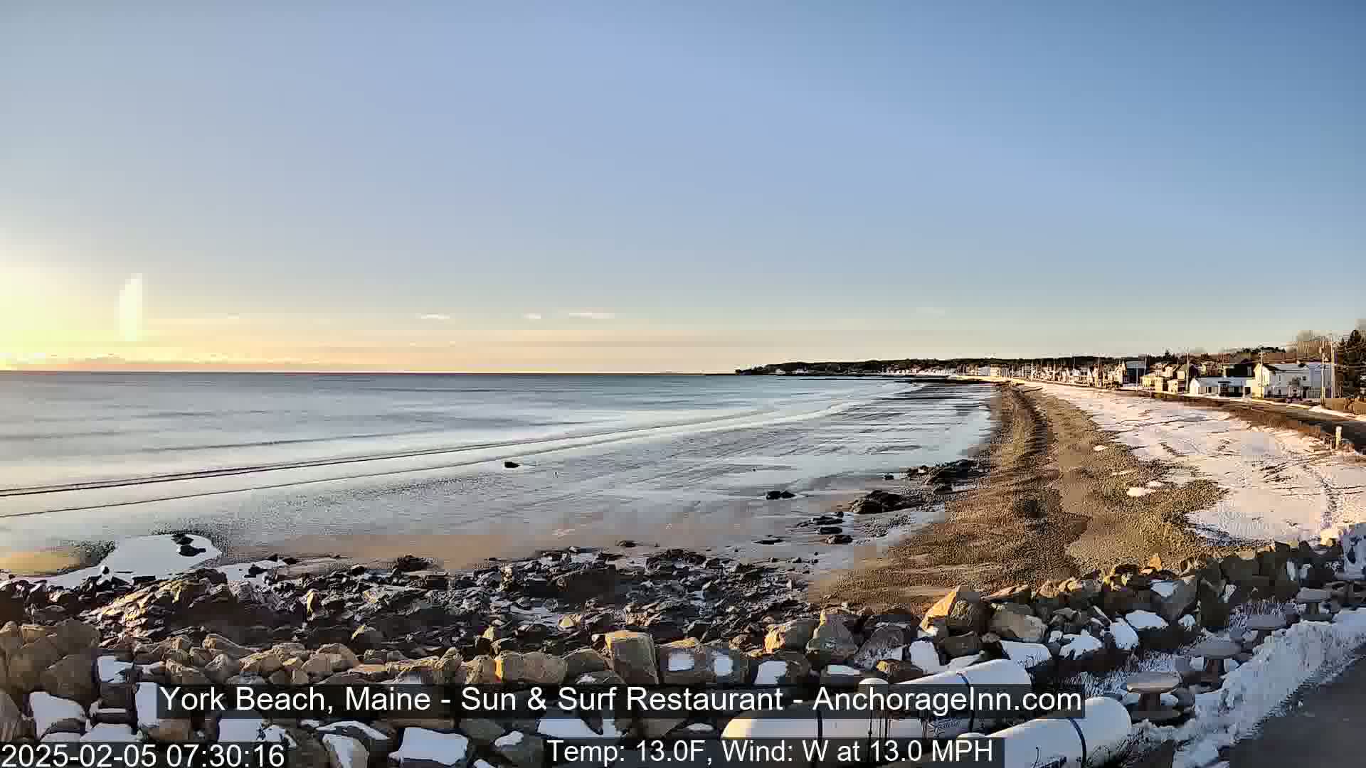 A long sandy beach with some snow and ice patches stretches along a calm ocean under a clear pale blue sky at sunrise.