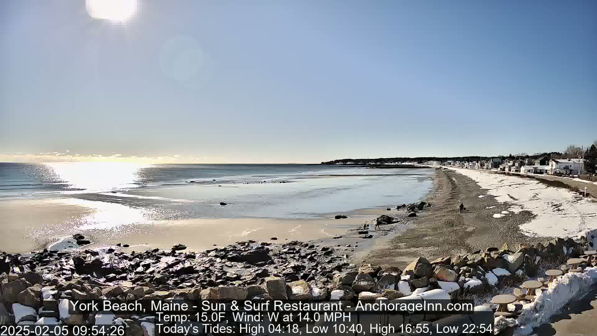 A sunny, cold day at a beach with patches of snow on the sand and rocks, overlooking a calm ocean with a small town in the distance.