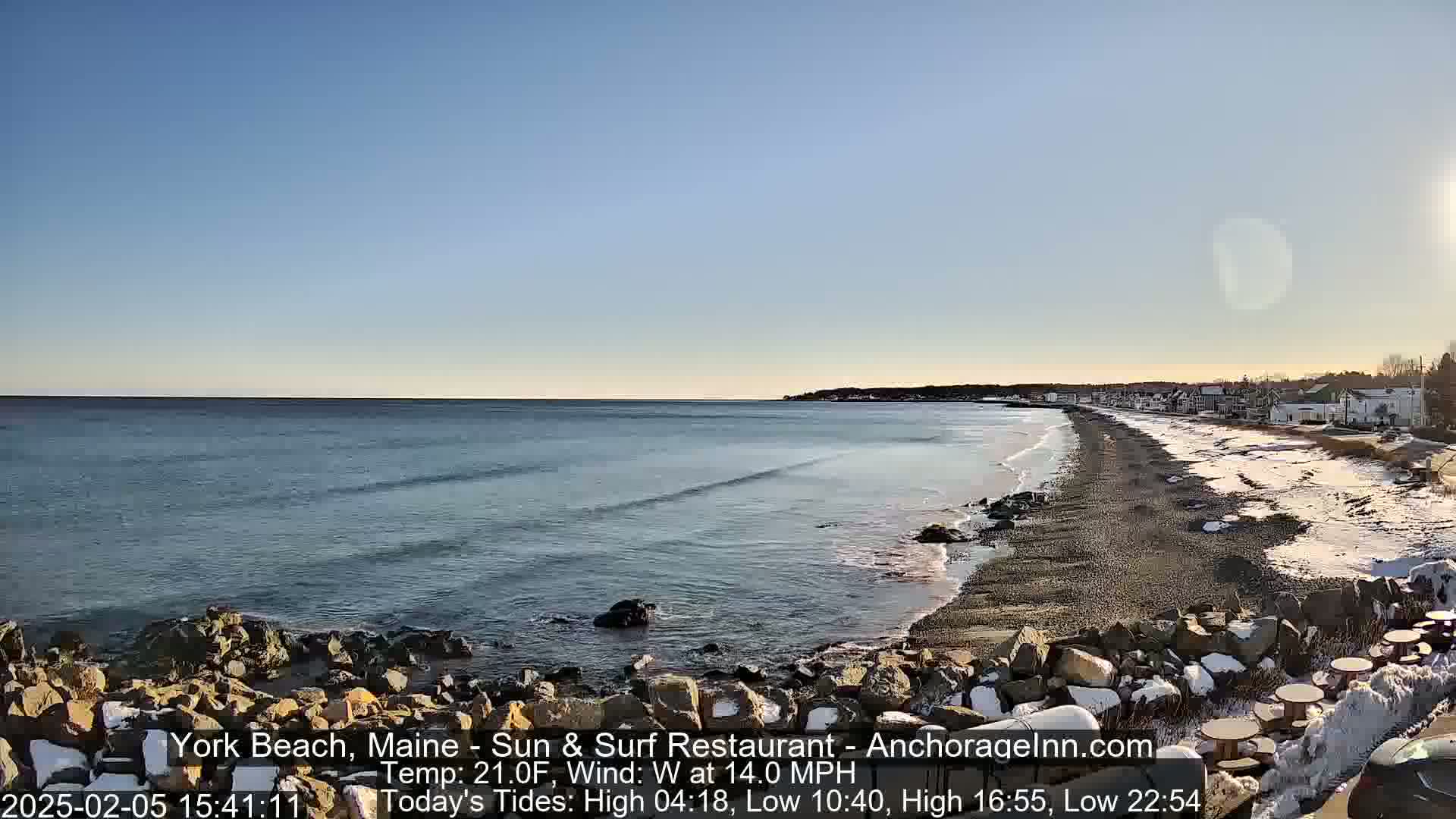 A partly sunny day shows a beach with a rocky shoreline and calm ocean waves, partially covered in snow.