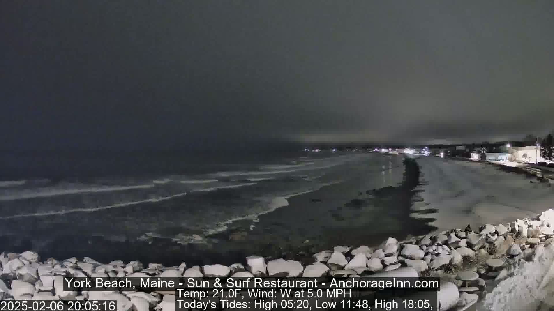 A dark, nighttime beach scene shows a rocky shoreline and gently lapping waves under a cloudy sky.