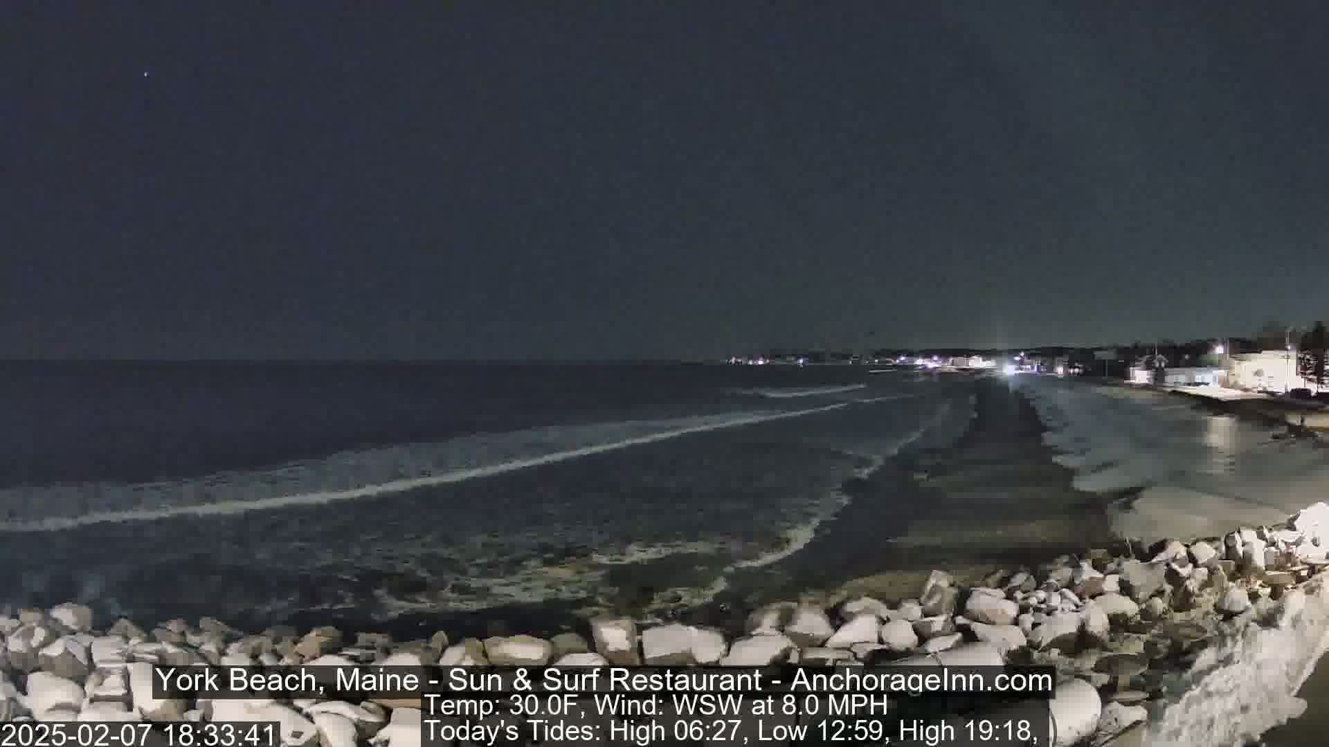 A nighttime view of a rocky shoreline with dark, gently lapping waves under a clear, dark sky.