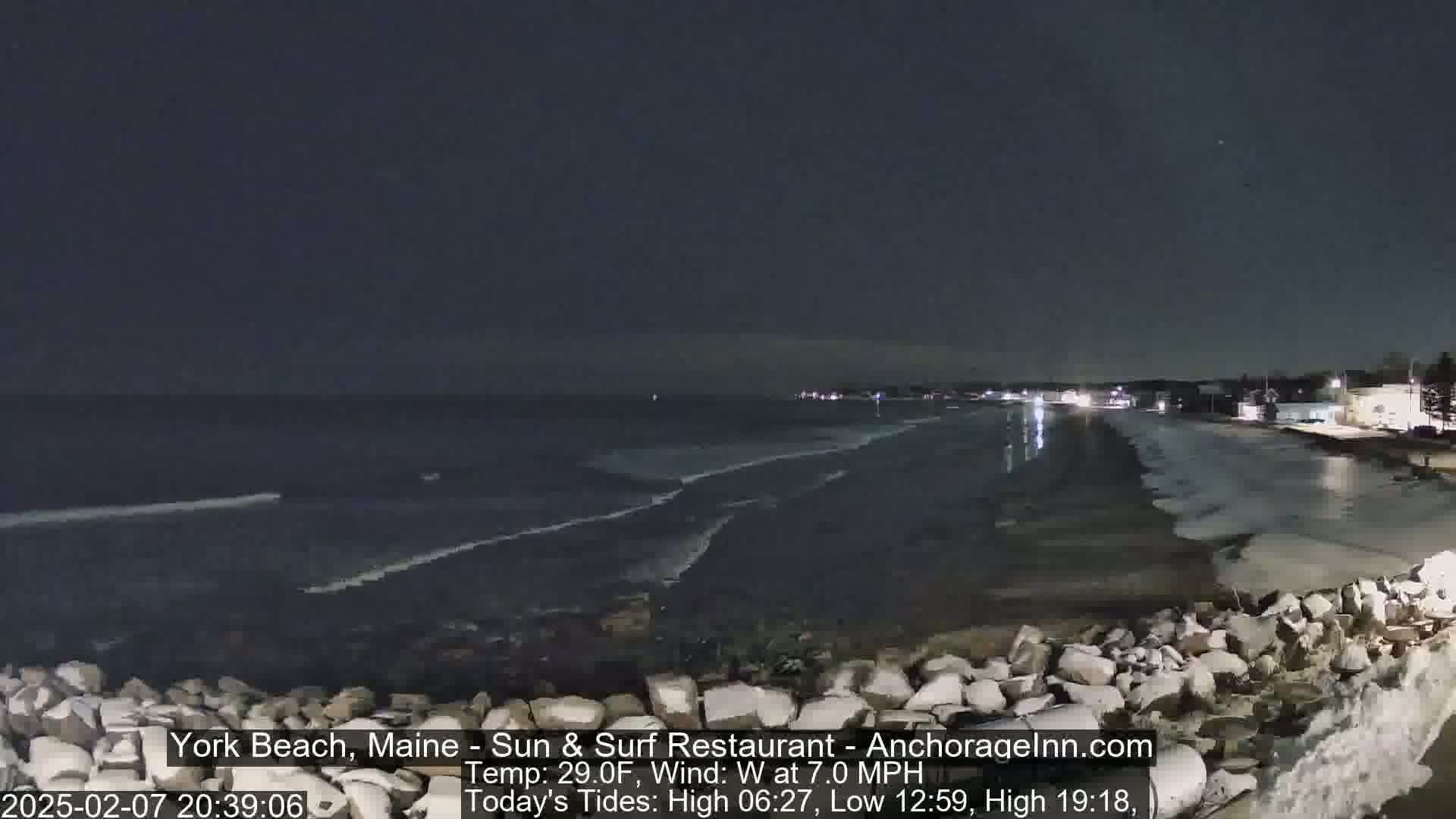 A dark nighttime beach scene shows a rocky shoreline, calm waves lapping the shore, and lights from buildings in the distance under a clear sky.