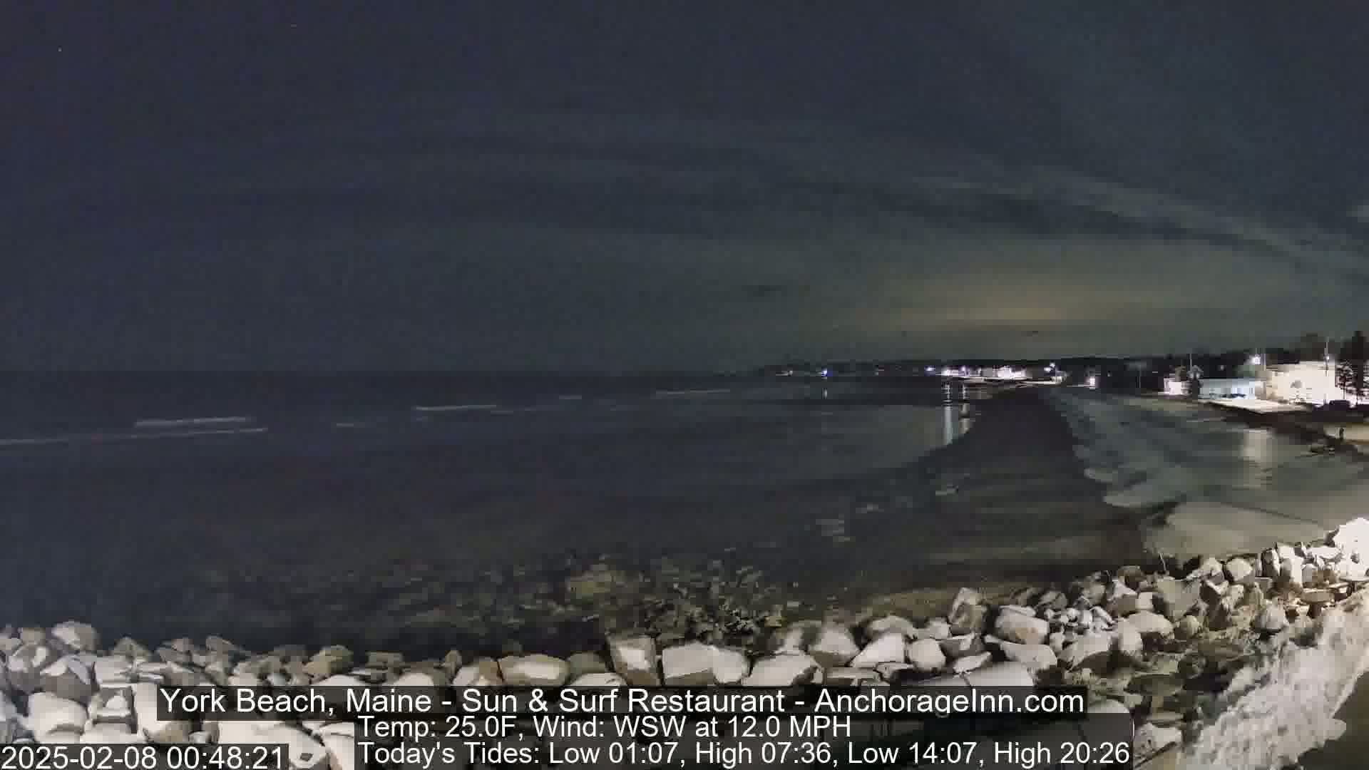 A nighttime, dark view of a rocky shoreline with a calm ocean under a mostly clear sky.