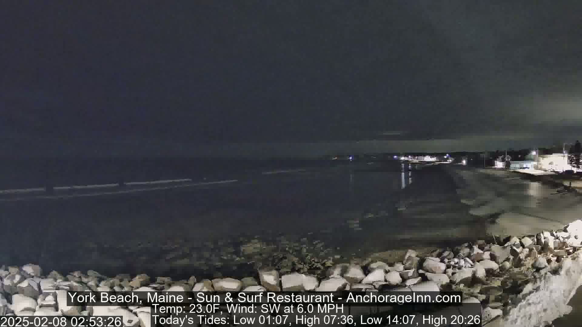 A nighttime view of a rocky shoreline and calm ocean under a dark, cloudy sky.