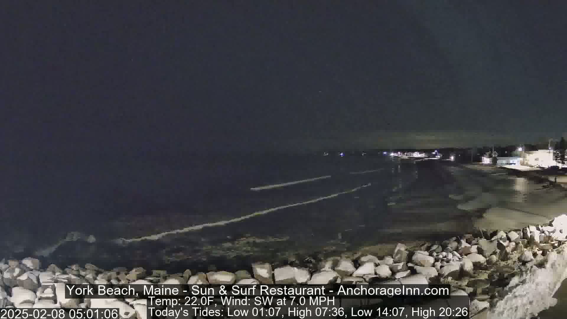 A dark, nighttime beach scene shows a rocky shoreline with gentle waves lapping under a clear, dark sky.