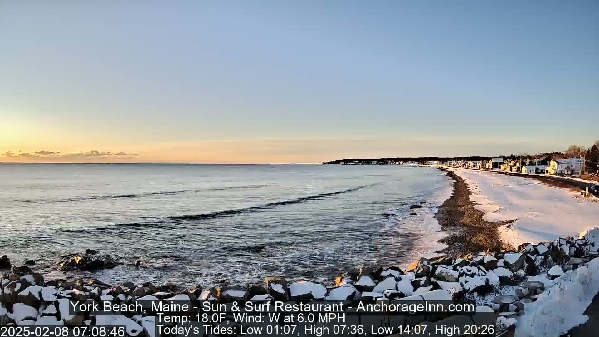 A snow-dusted beach with calm ocean waves stretches along a coastline of houses under a clear, sunrise sky.