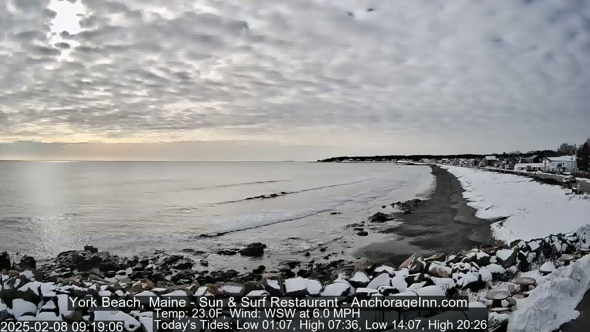 A snow-covered rocky shoreline meets a calm, gray ocean under a cloudy sky.