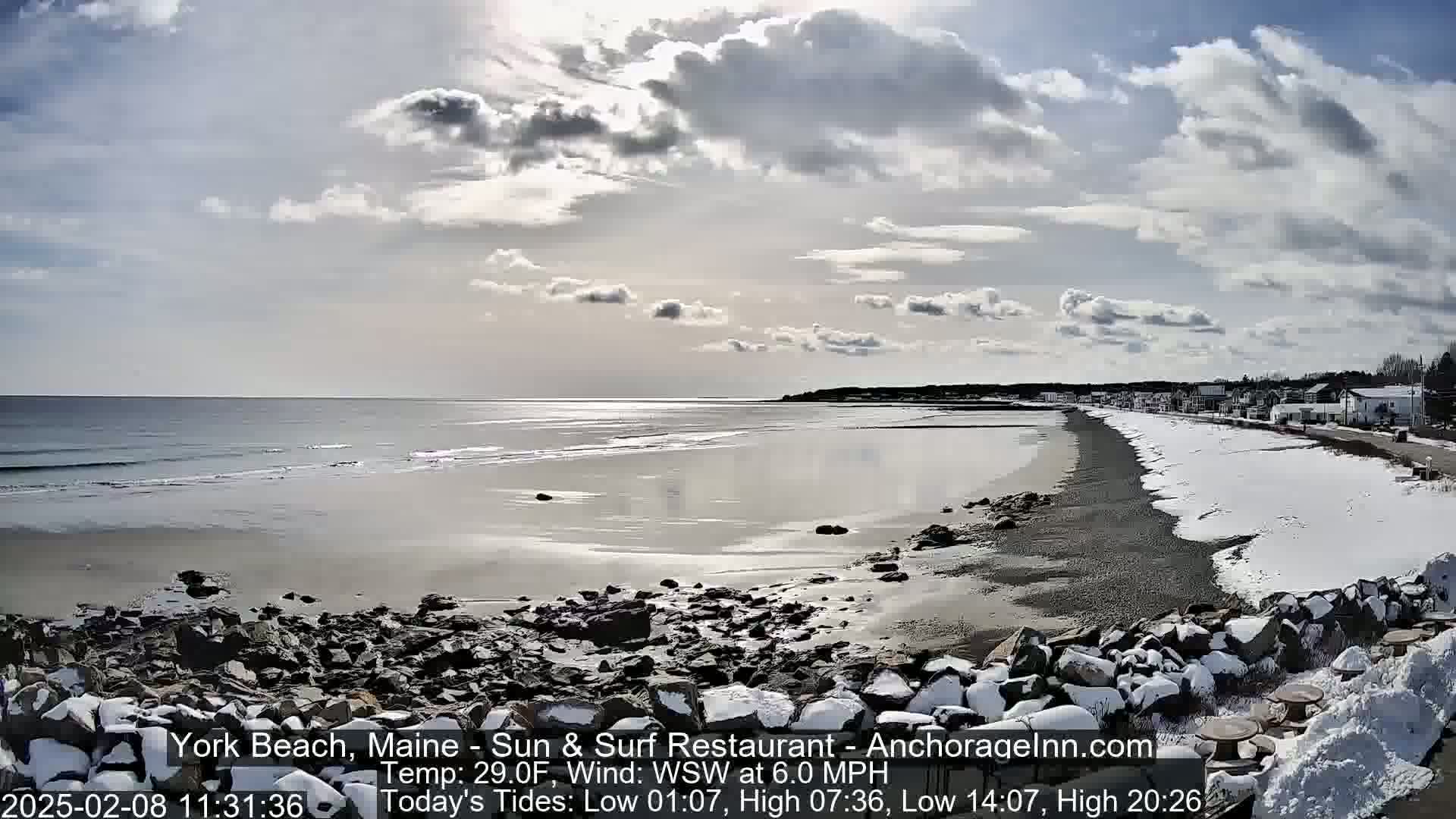 A partly cloudy day shows a partially snow-covered beach with dark rocks and calm ocean waves.