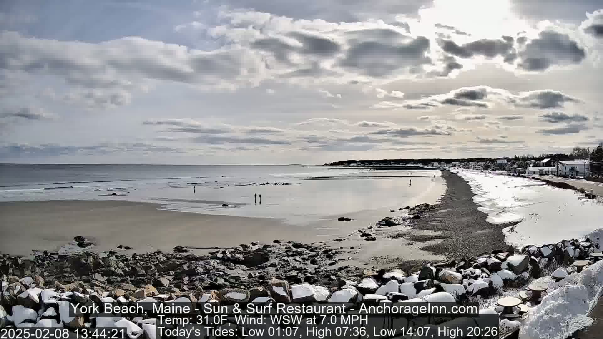 A long, sandy beach with some snow and ice stretches toward a small town under a partly cloudy sky.