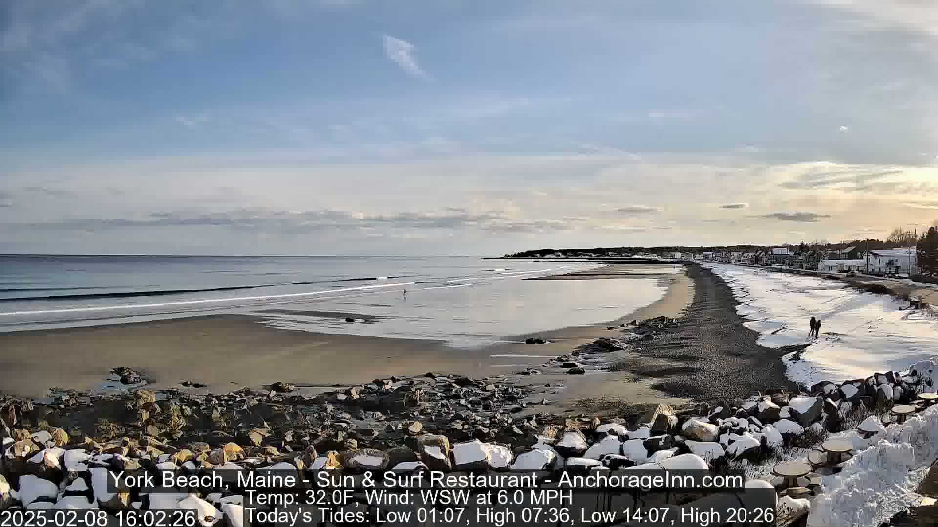A sandy beach with some snow on the shore and rocks, partially covered in snow, extends to a calm ocean under a mostly clear sky.