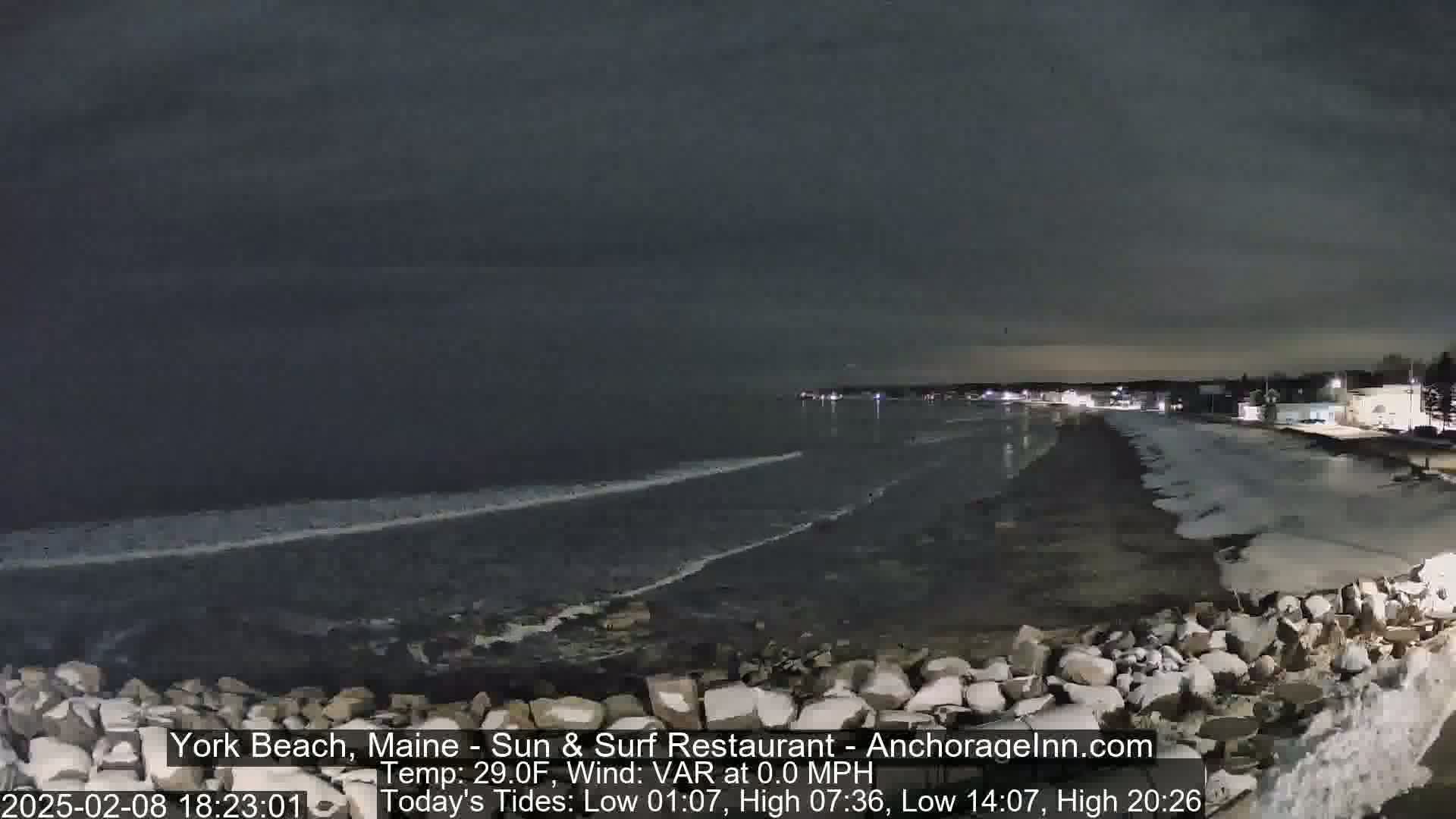 A dark, nighttime view of a rocky beach with calm ocean waves under a cloudy sky.