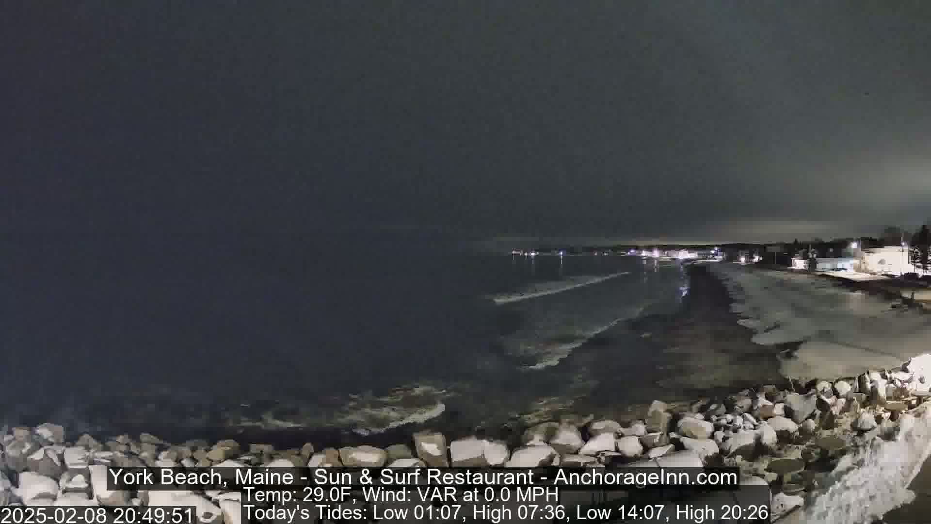 A nighttime view of a rocky shoreline with a dark, calm ocean under a cloudy sky.