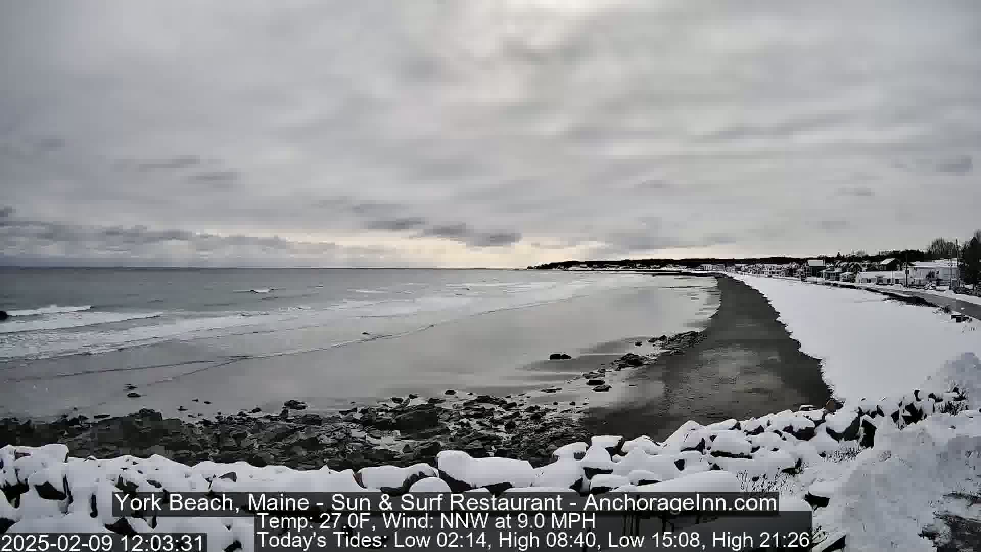 A snow-covered rocky shoreline meets a gray ocean with small waves under an overcast sky.