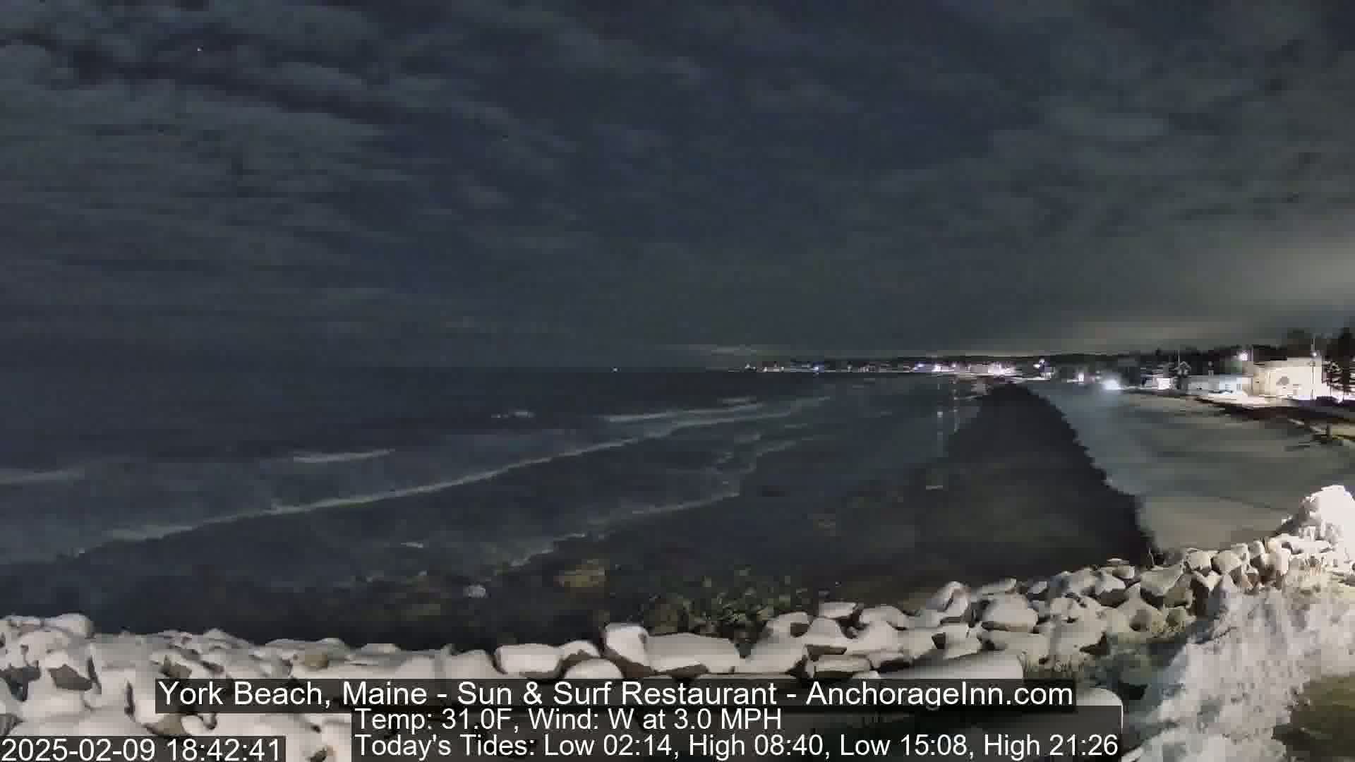 A nighttime view of a dark ocean with gentle waves lapping against a rocky shoreline under a cloudy sky.