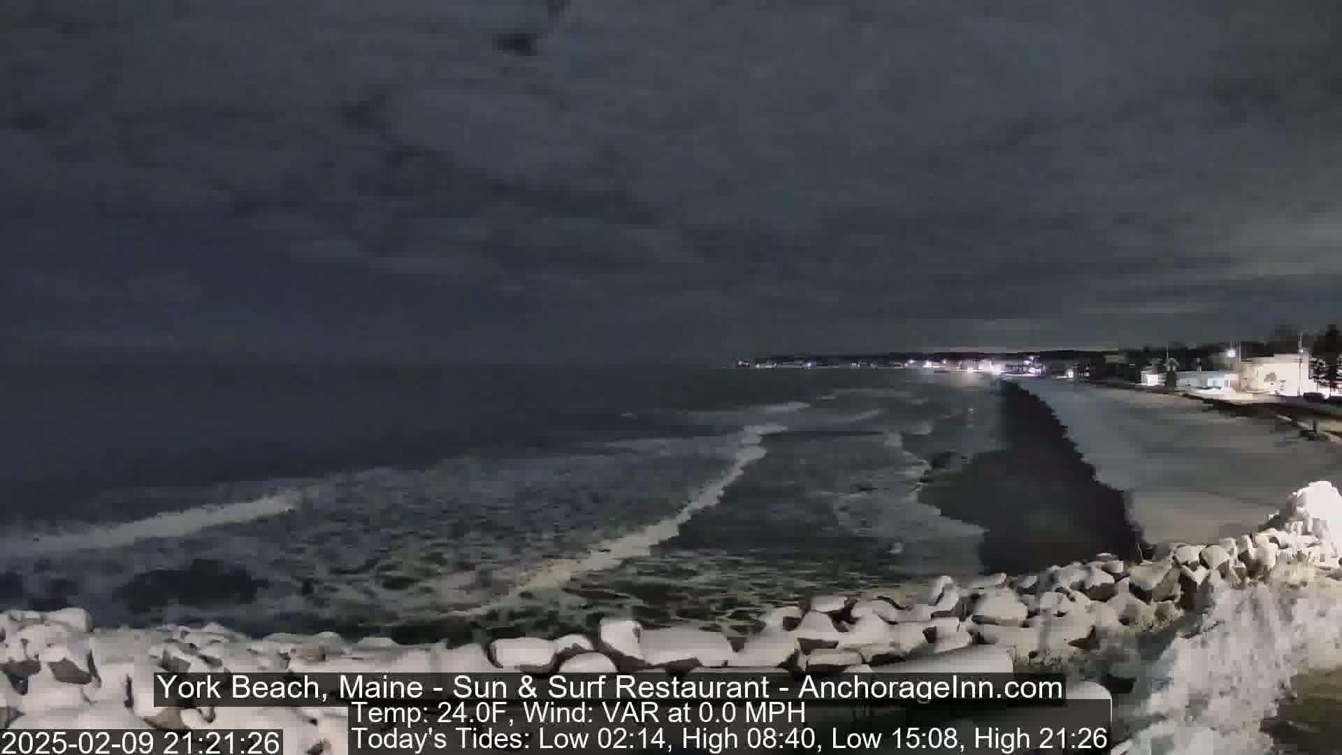 A dark, nighttime coastal scene shows a snow-dusted rocky shoreline with small waves lapping against it under a cloudy sky.