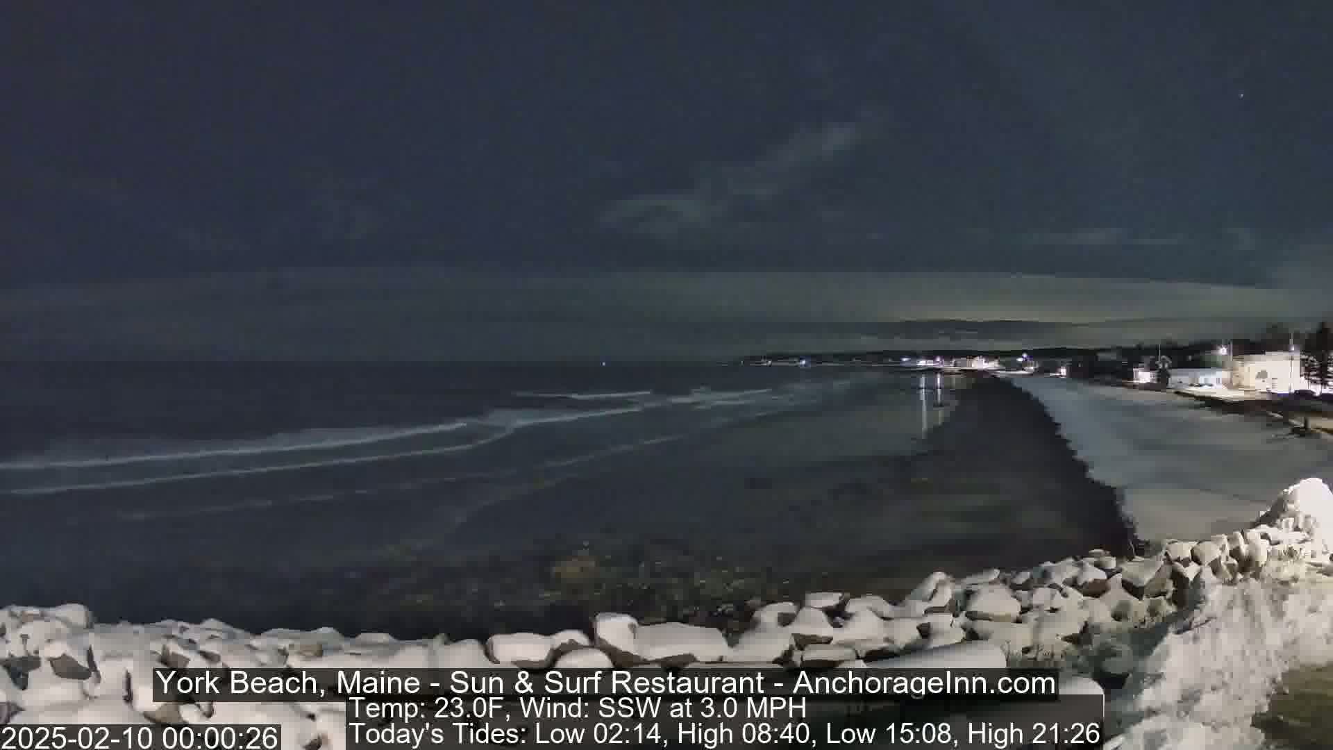 A snow-dusted rocky shoreline at night overlooks a dark ocean with small waves under a mostly clear sky.