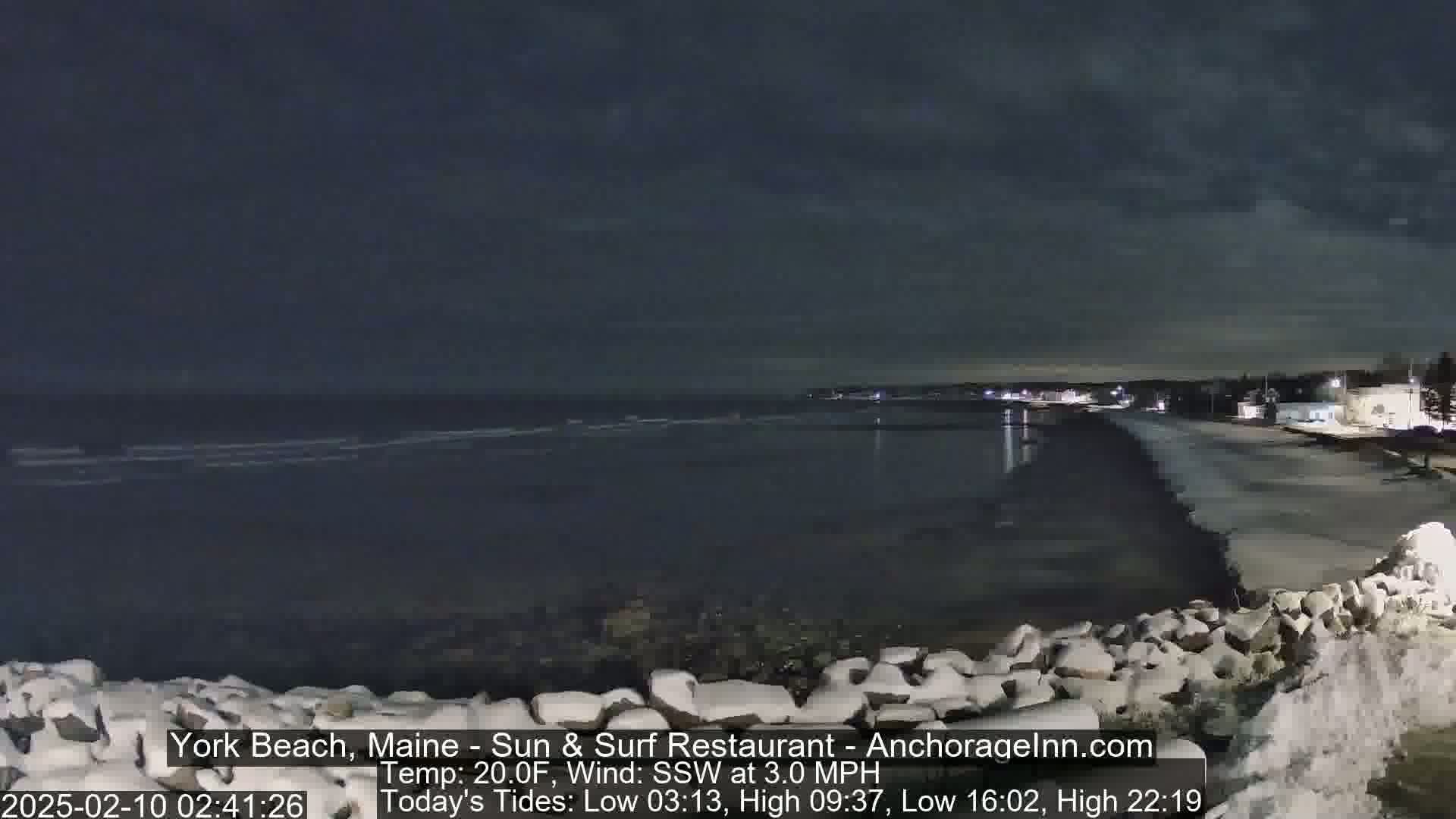 A dark, nighttime beach scene shows a calm ocean with small waves gently lapping the shore under a mostly clear sky, with a temperature of 20°F.