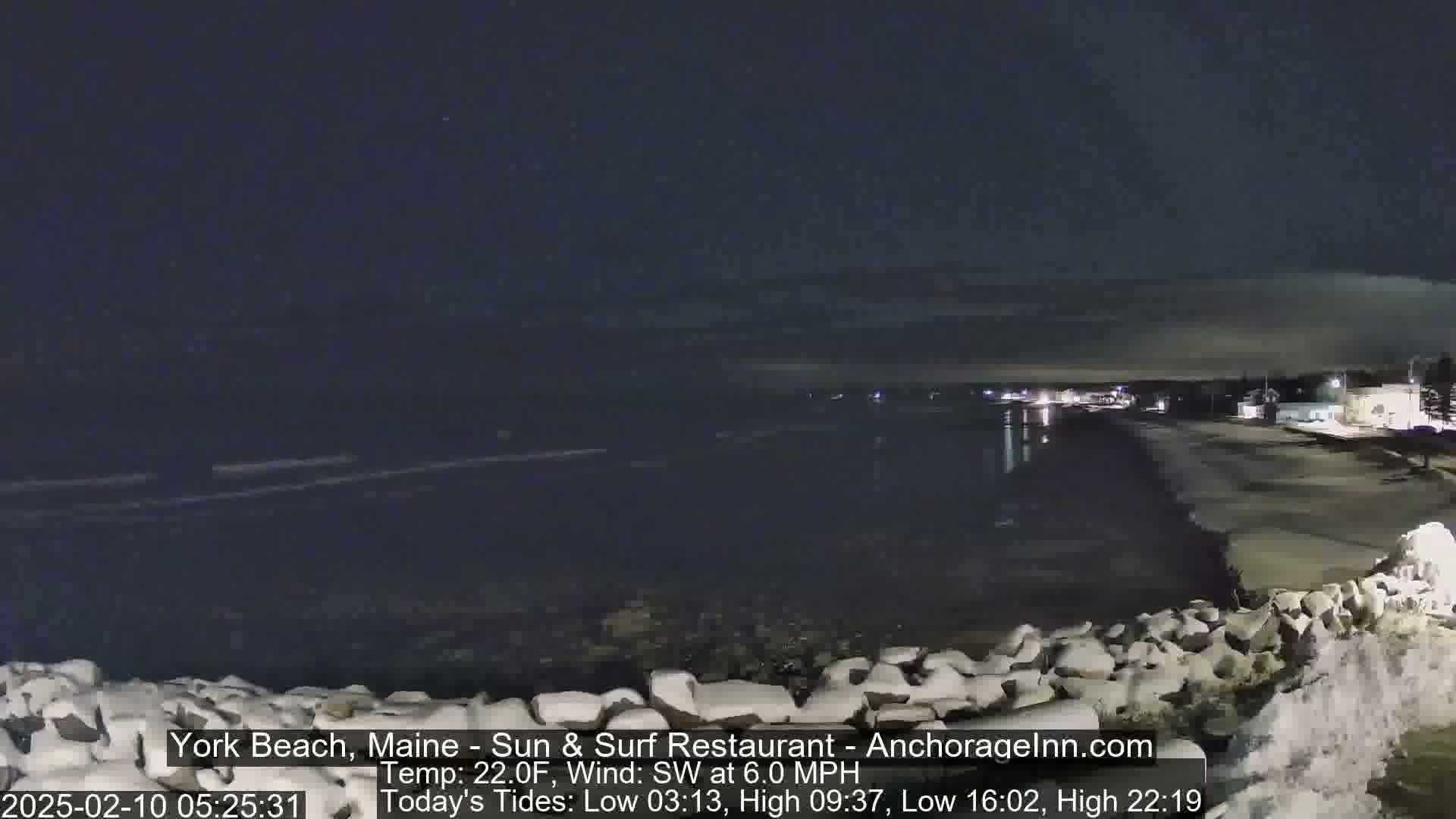 A nighttime view of a dark, calm ocean beach with a few lights visible in the distance under a mostly clear sky.