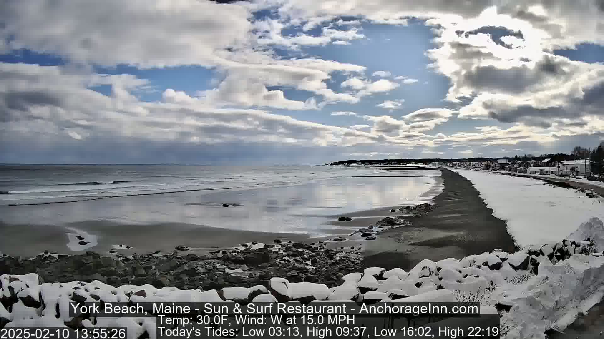 A partly cloudy, windy day at a snow-covered beach shows a calm ocean with small waves, a dark sandy shore, and a town in the distance.