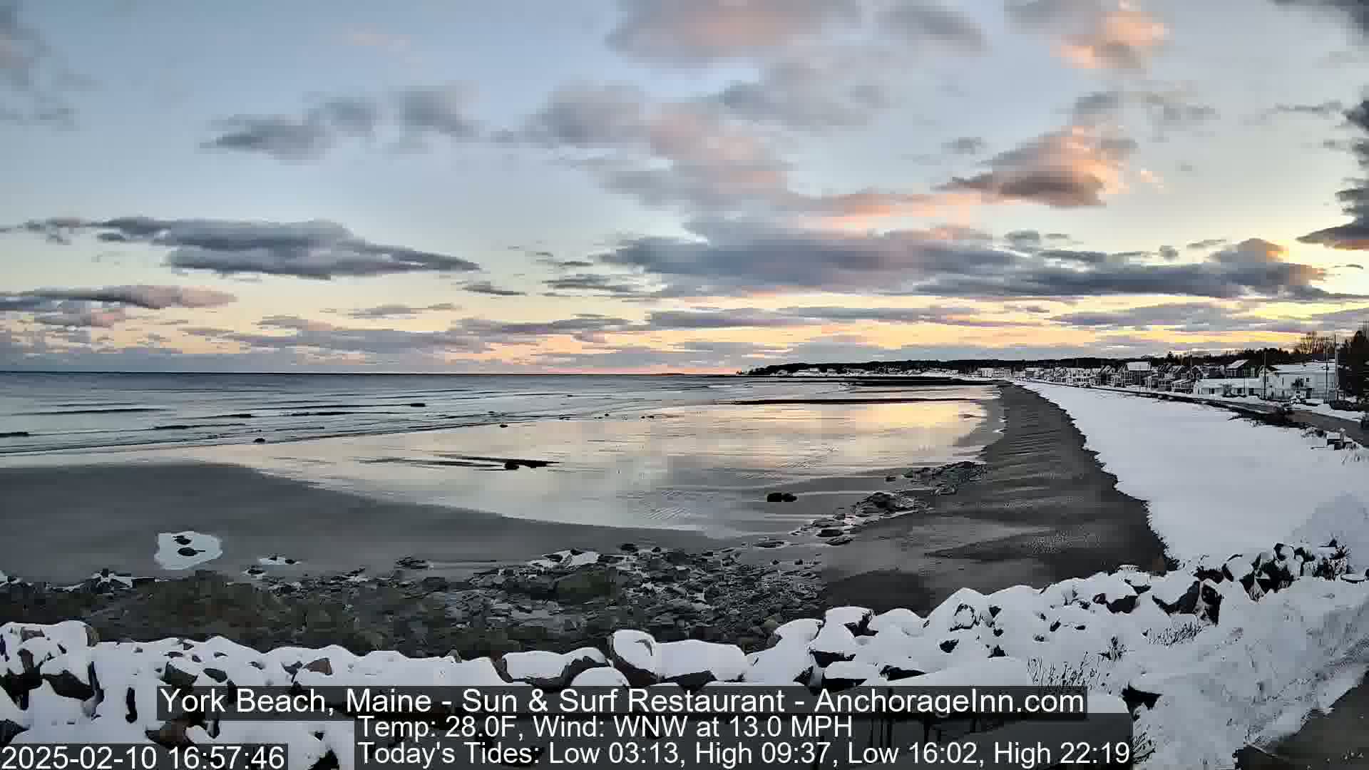 A snow-covered beach at sunset, with a calm ocean reflecting the pale pink and orange sky, and a town visible in the distance.