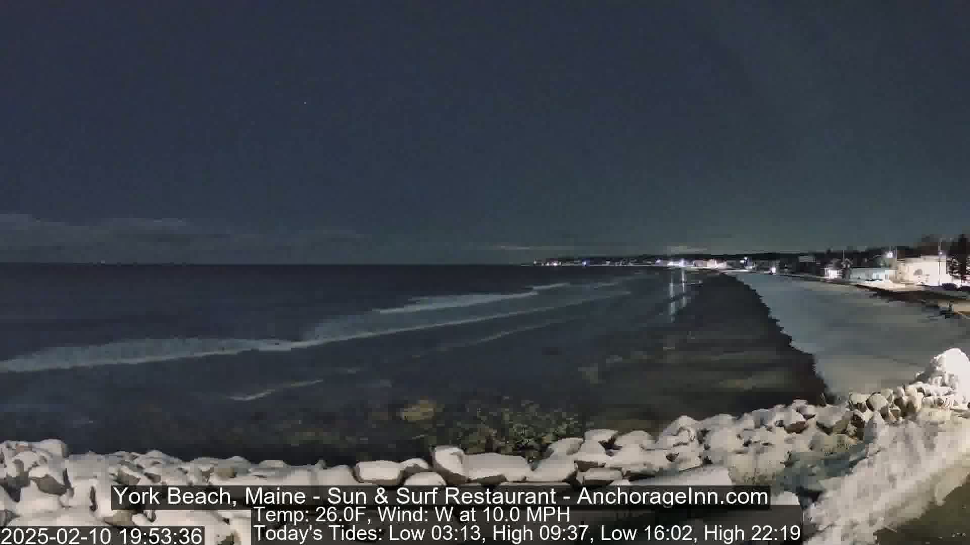A dark, nighttime beach scene shows a partially frozen, snow-covered shoreline with a calm ocean under a clear, starry sky, and a slight wind.