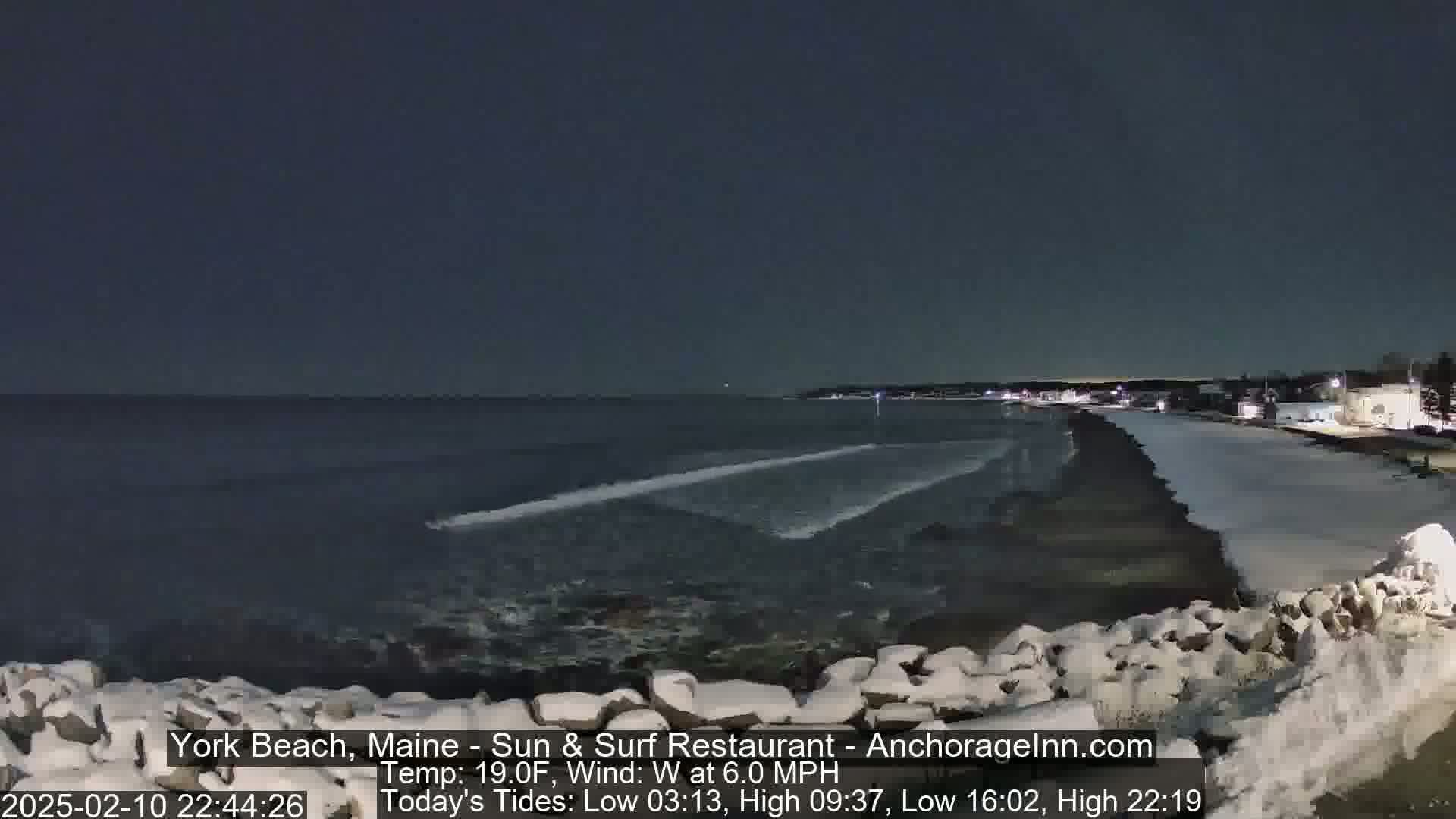 A dark, nighttime beach scene shows a partially frozen, snow-covered shoreline with gentle waves lapping at the shore under a clear, dark sky.