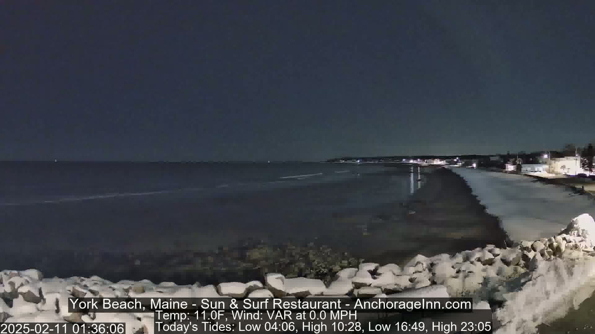 A dark, nighttime coastal scene shows a calm, dark ocean, a snow-covered beach, and a partially snow-covered rocky wall in the foreground under a clear, dark sky.