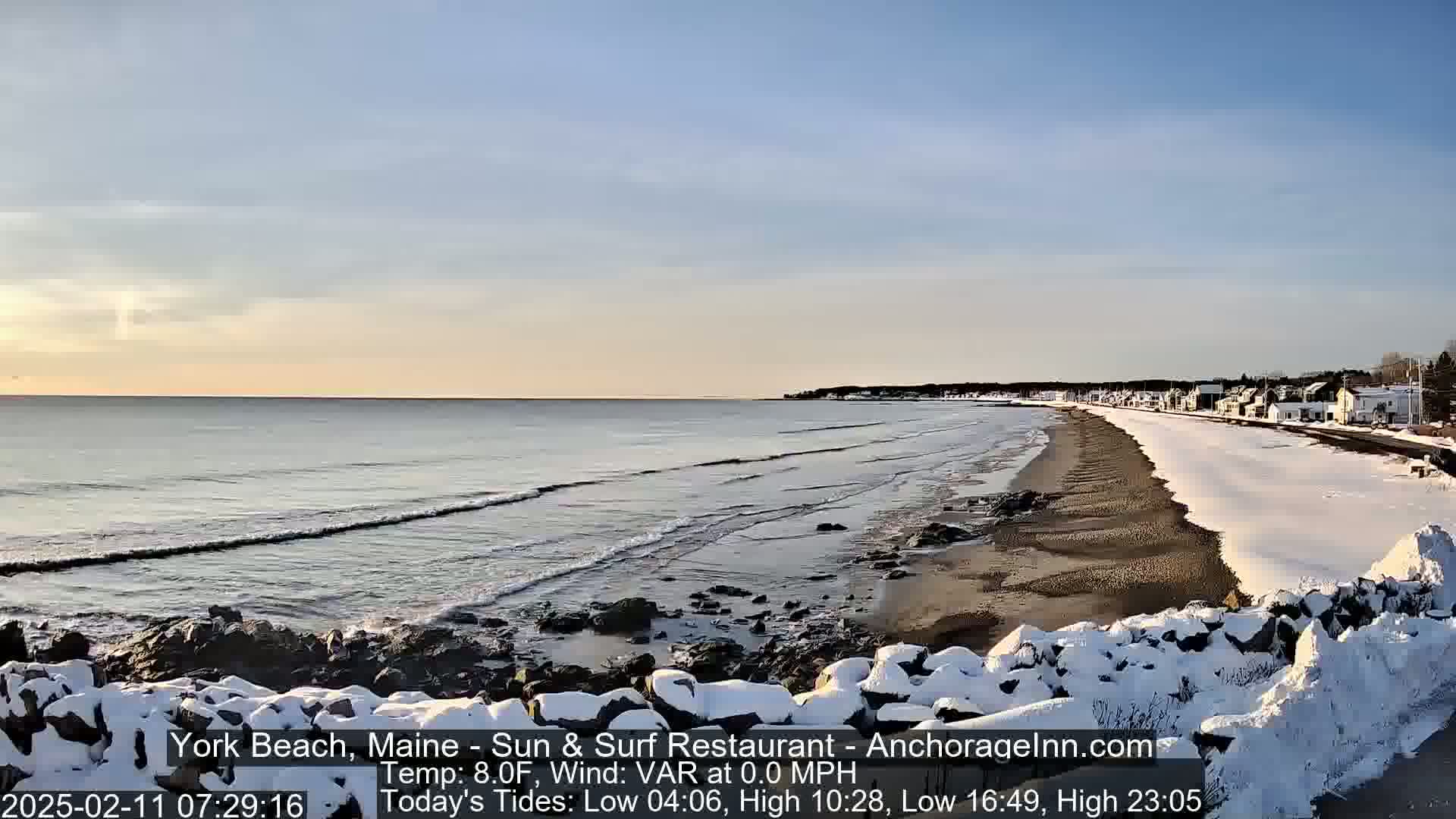 A snow-covered beach with calm waves rolls onto the shore under a clear, bright sky.