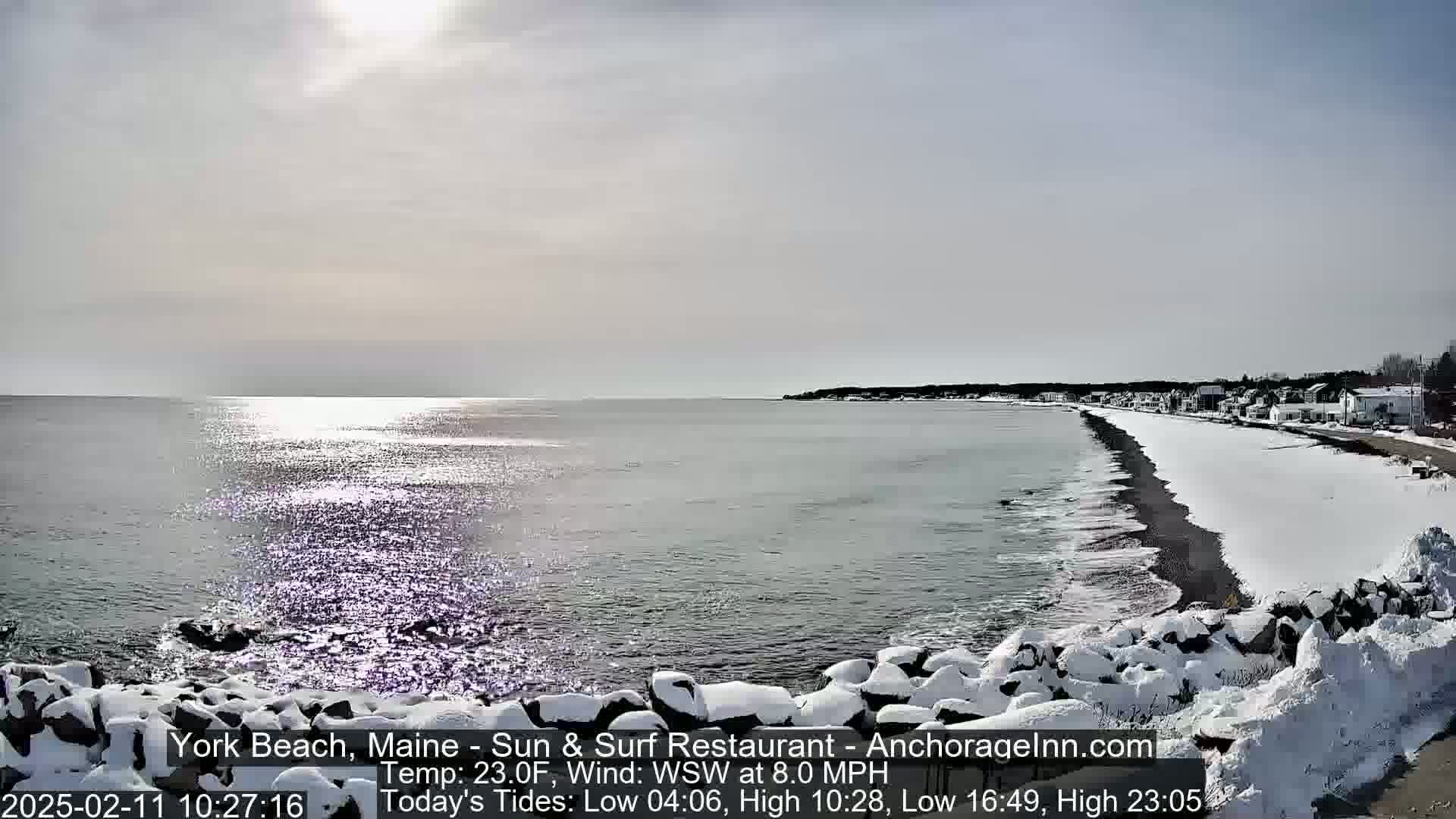 A snow-covered beach and shoreline with a calm, sunlit ocean in the background.