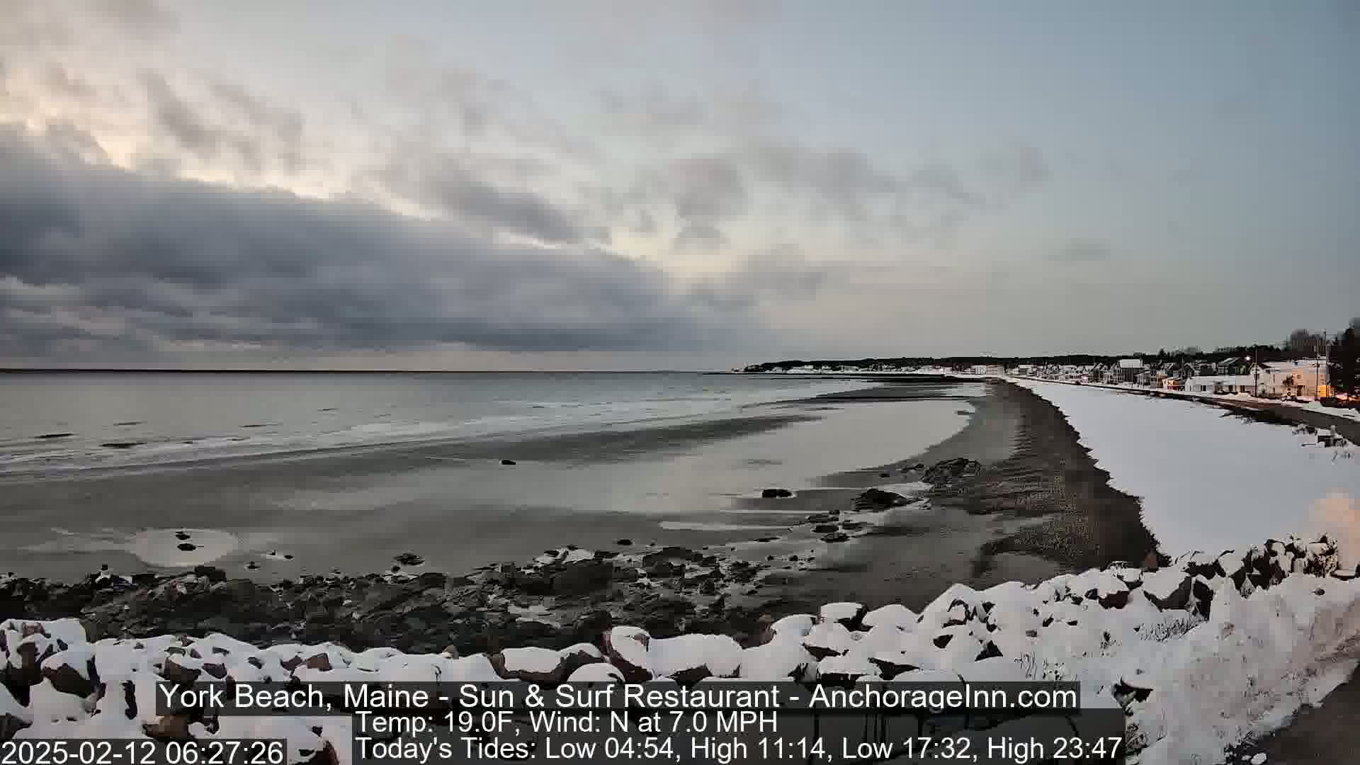 A snow-dusted beach stretches along a calm, gray ocean under a cloudy sky.
