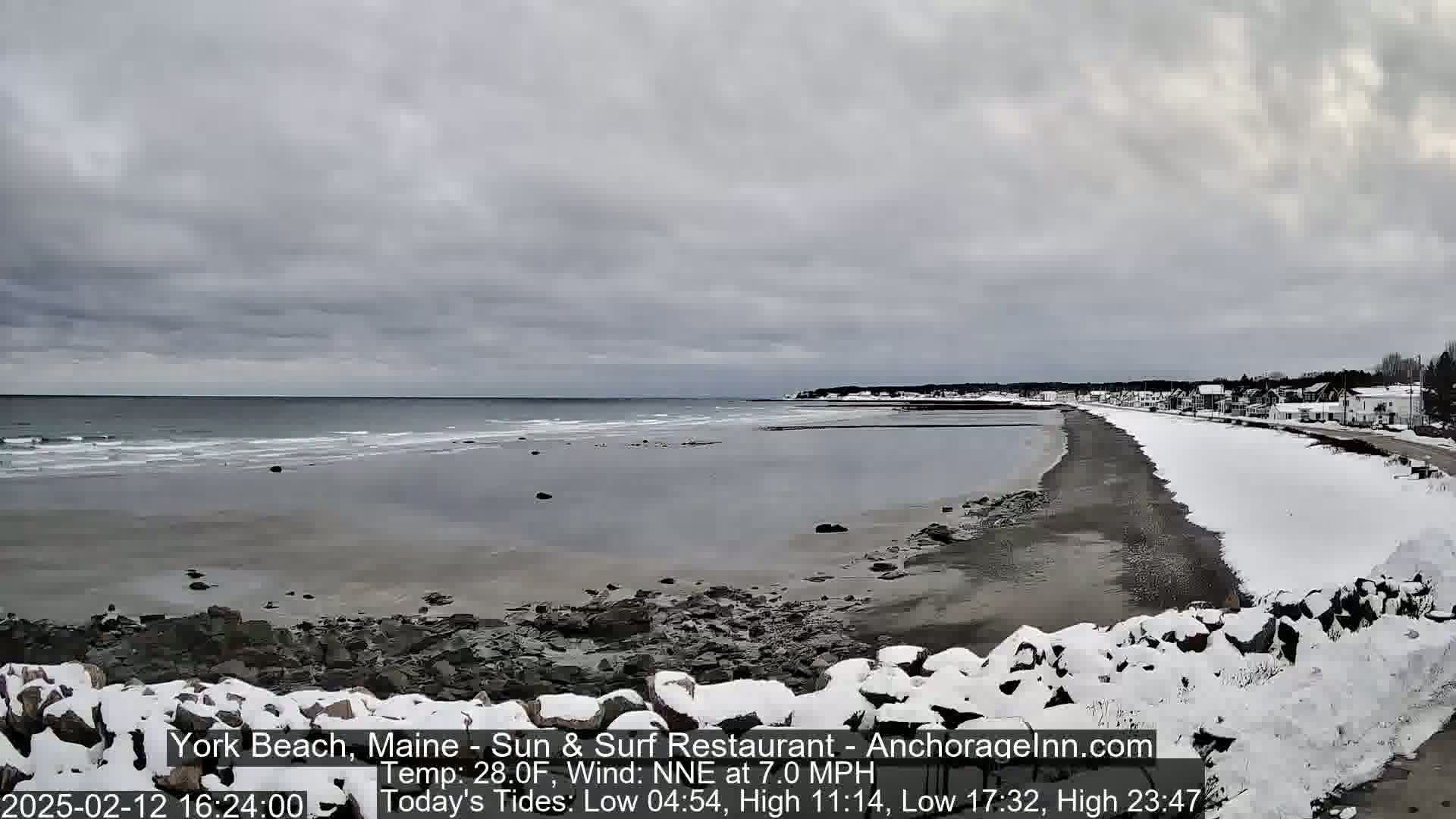 A snow-covered rocky shoreline stretches alongside a grey, overcast ocean with small waves under a cloudy sky.