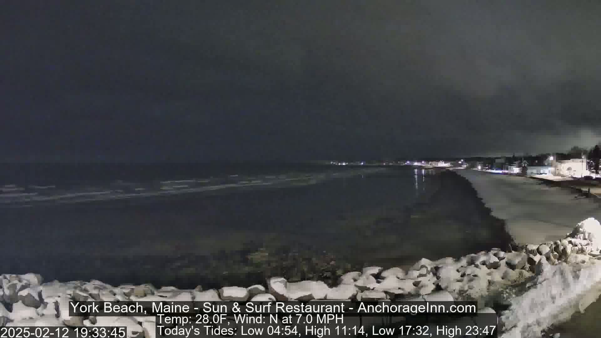A dark, nighttime beach scene with a partially snow-covered rocky shoreline, calm ocean waves, and a dimly lit town in the background under a cloudy sky.