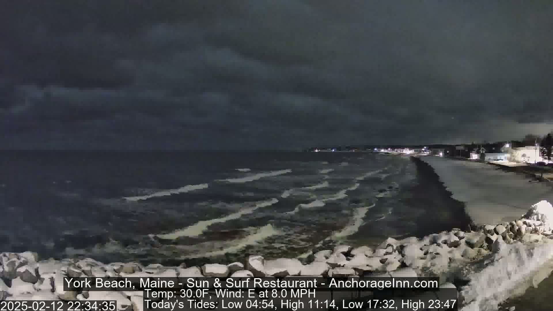 A nighttime view of a snow-covered rocky shoreline with dark, choppy waves under a cloudy sky.