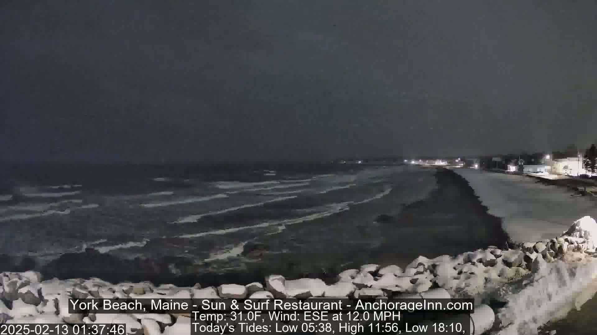 A snow-covered rocky shoreline at night shows a dark, choppy sea under a dark sky.