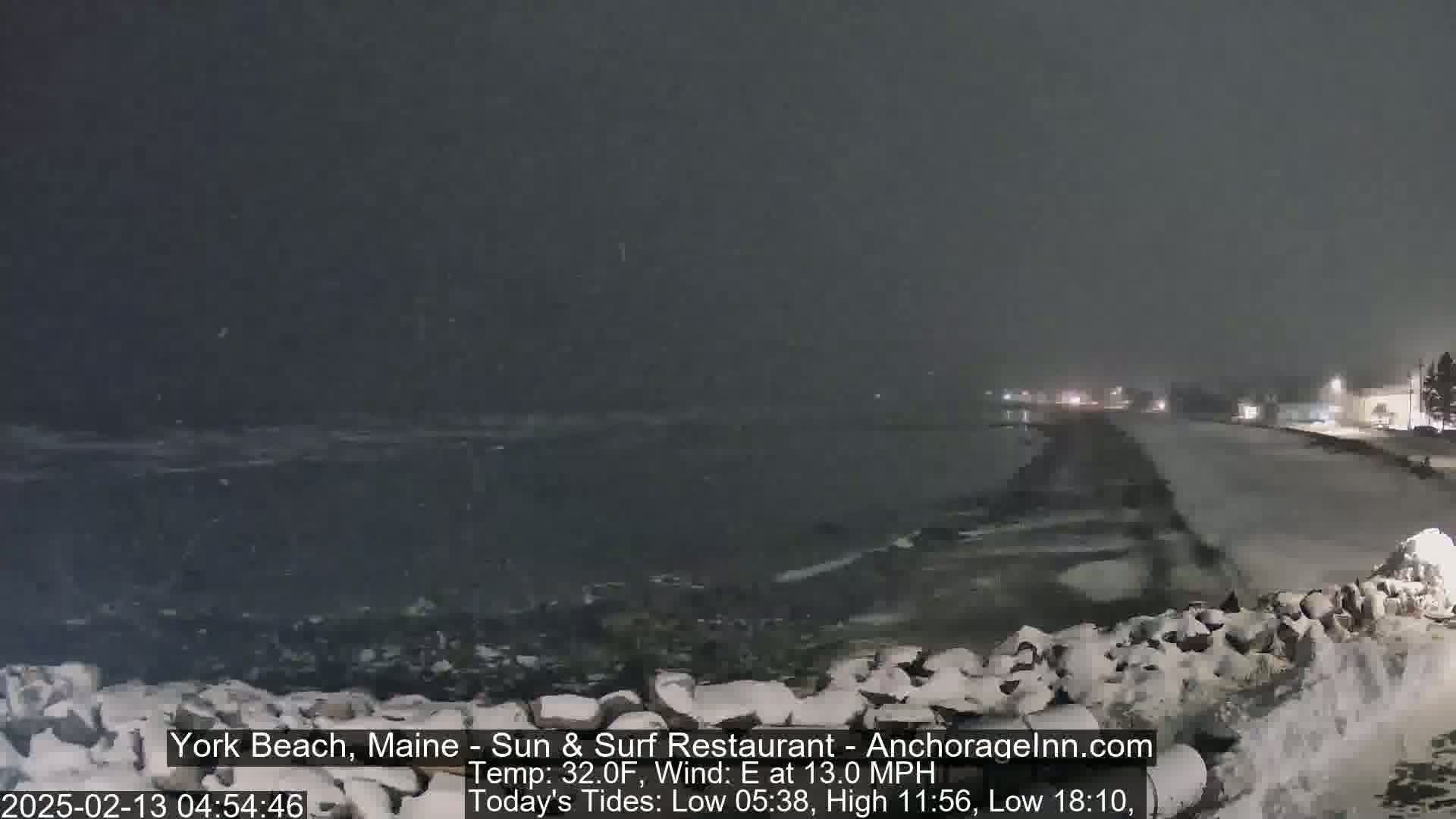 A snow-covered shoreline at night, with dark, choppy water and a snow-covered road visible beyond a low stone wall in the heavy snowfall.