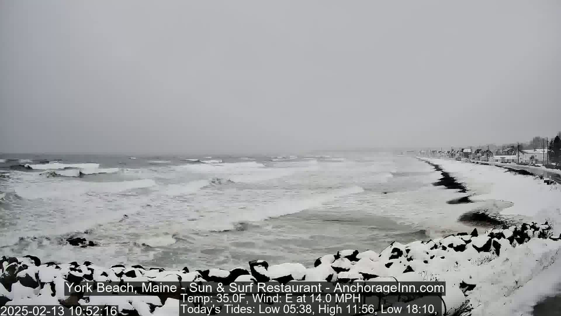 A snow-covered beach with rocky shoreline and waves crashing during a snowy, overcast day.