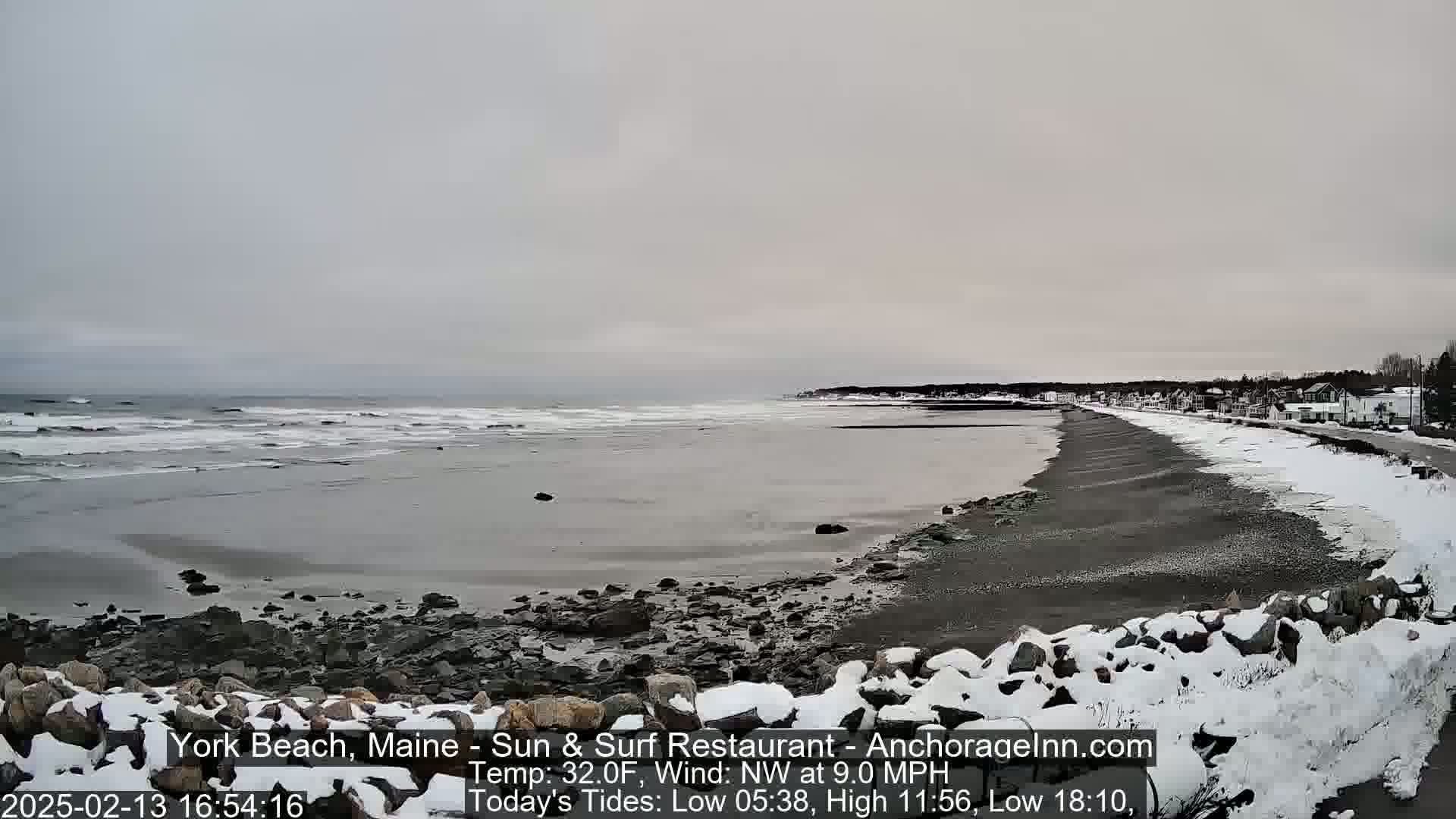 A snow-dusted beach stretches along a gray, overcast ocean under a cloudy sky with small waves.