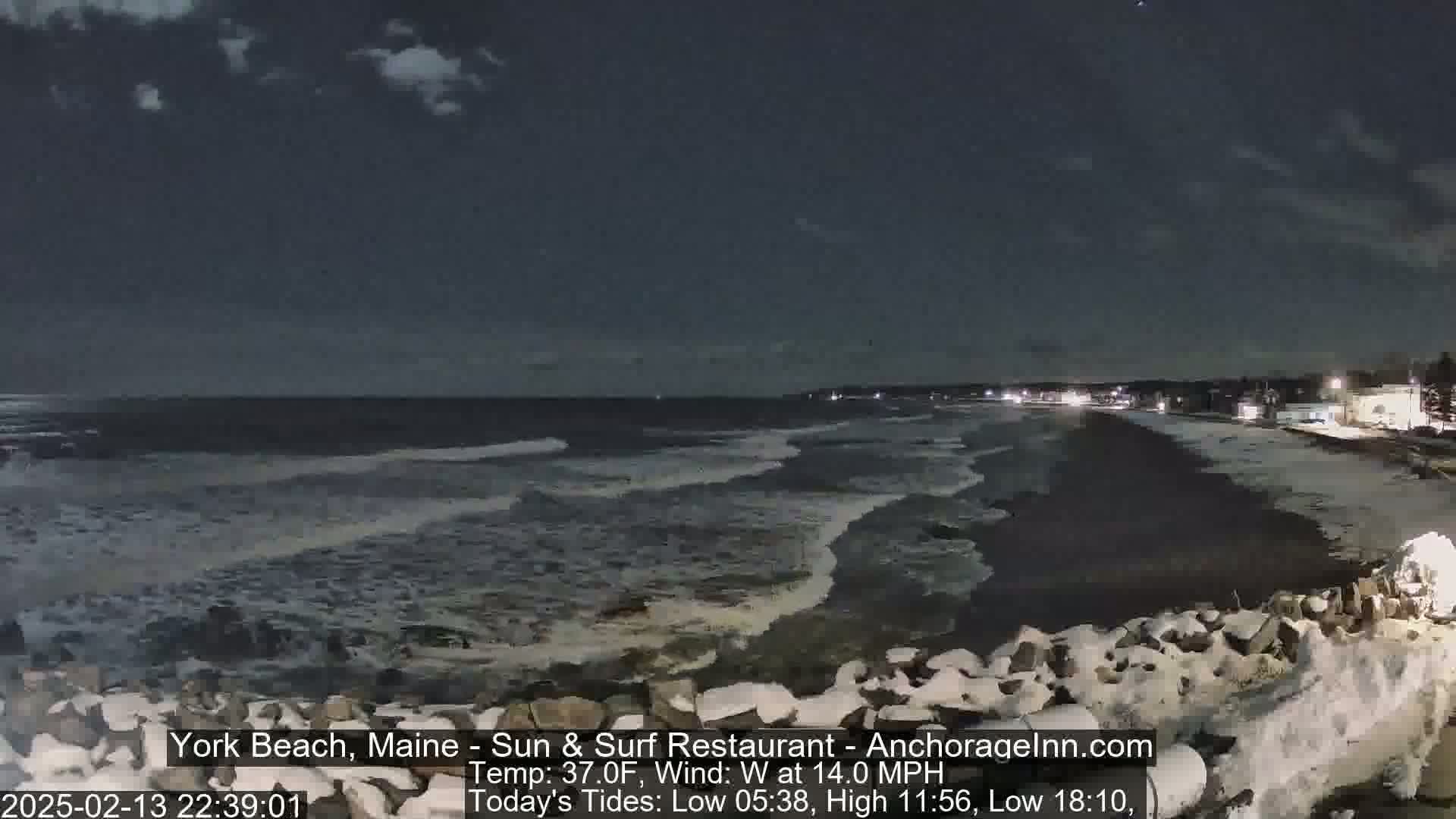 A nighttime view of a snow-dusted rocky shoreline and dark ocean waves under a partly cloudy sky.