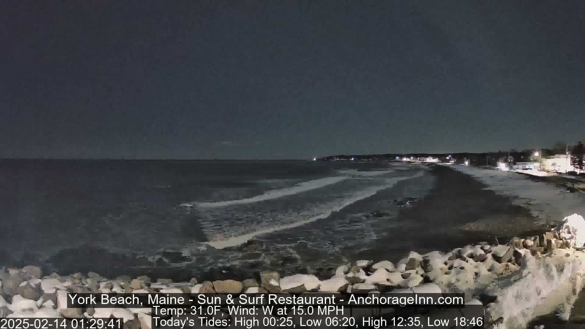 A nighttime beach scene shows dark, wavy water lapping against a snow-dusted rocky shoreline under a dark sky.
