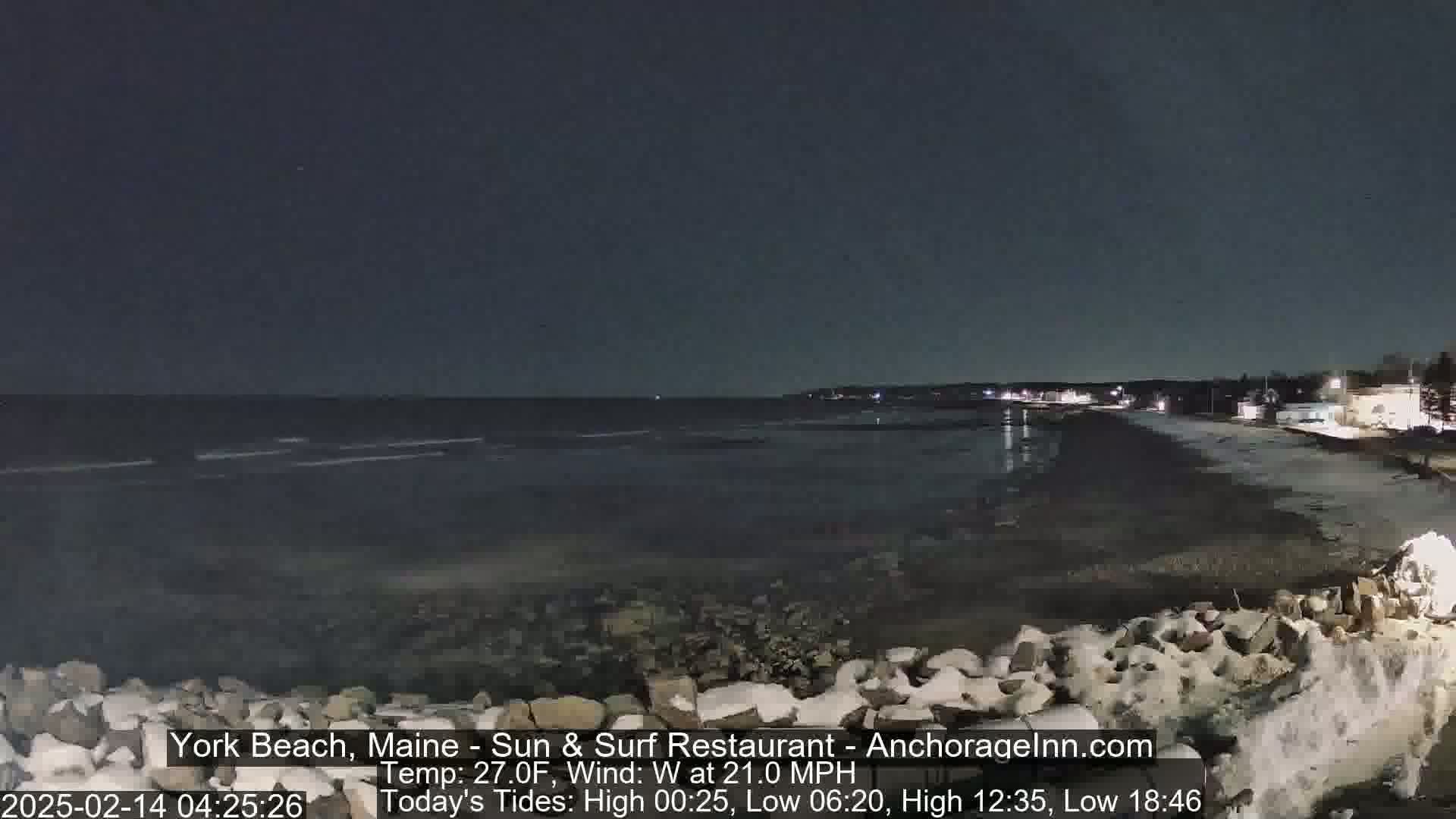 A nighttime view of a snow-dusted beach with dark, wavy water under a clear sky, a slight wind blowing.