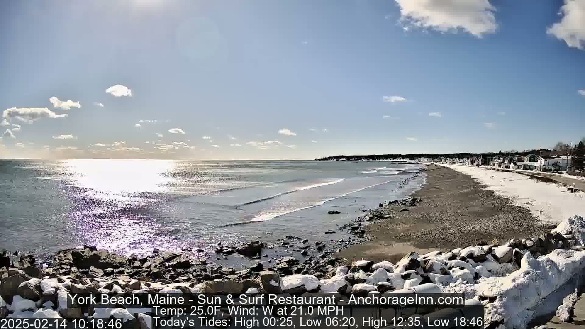 A sandy beach partially covered in snow stretches along a rocky shoreline, with a calm ocean reflecting sunlight under a partly cloudy sky.