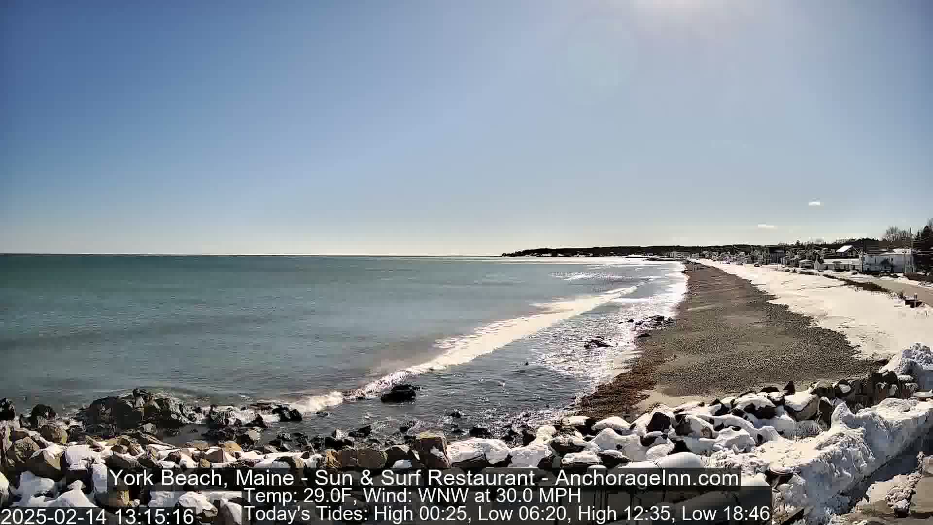 A sandy beach partially covered in snow stretches along a rocky coastline, with a calm ocean under a clear blue sky.